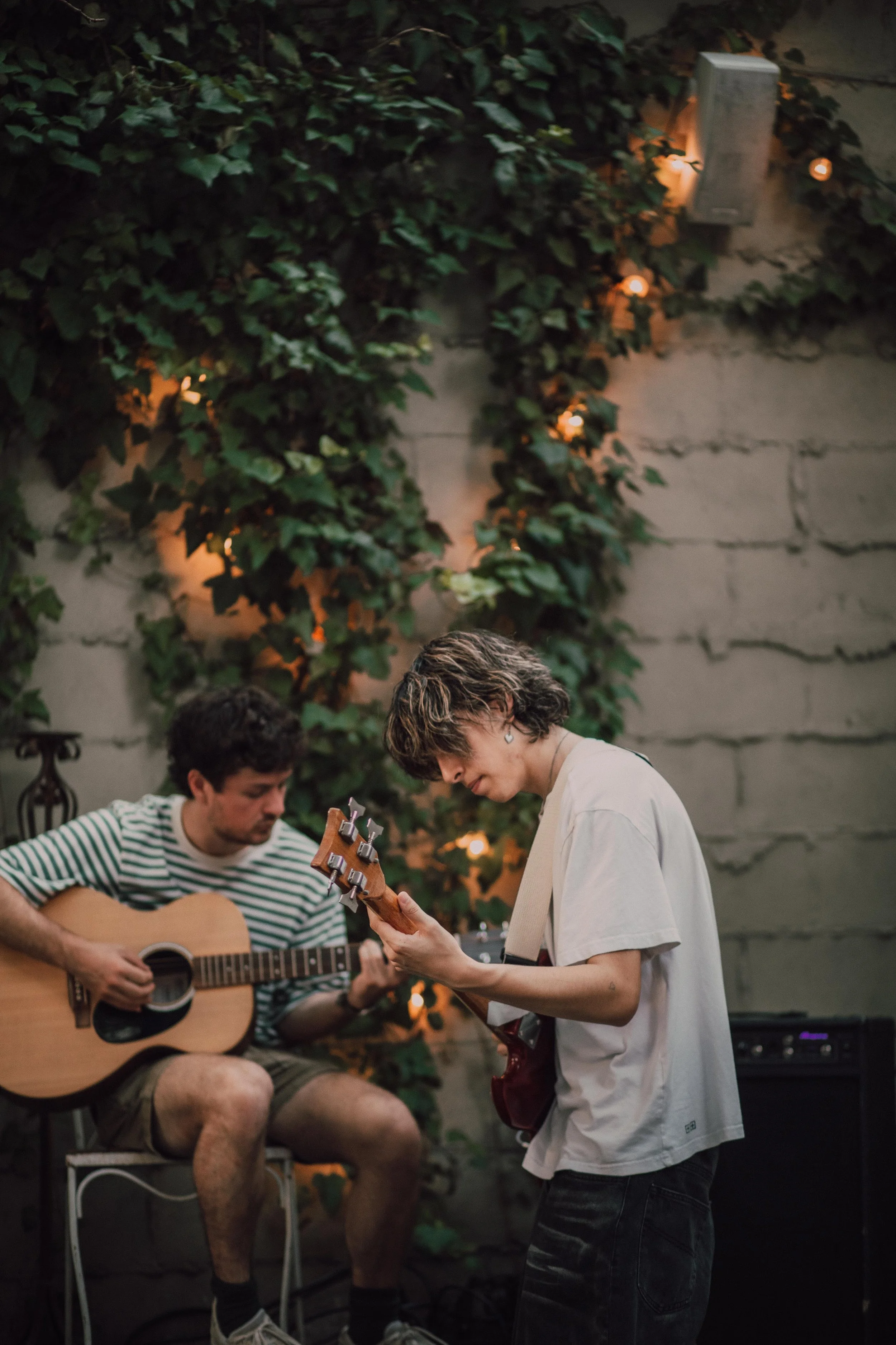 Two young adults playing guitars and singing together in a cozy indoor space decorated with string lights and greenery.