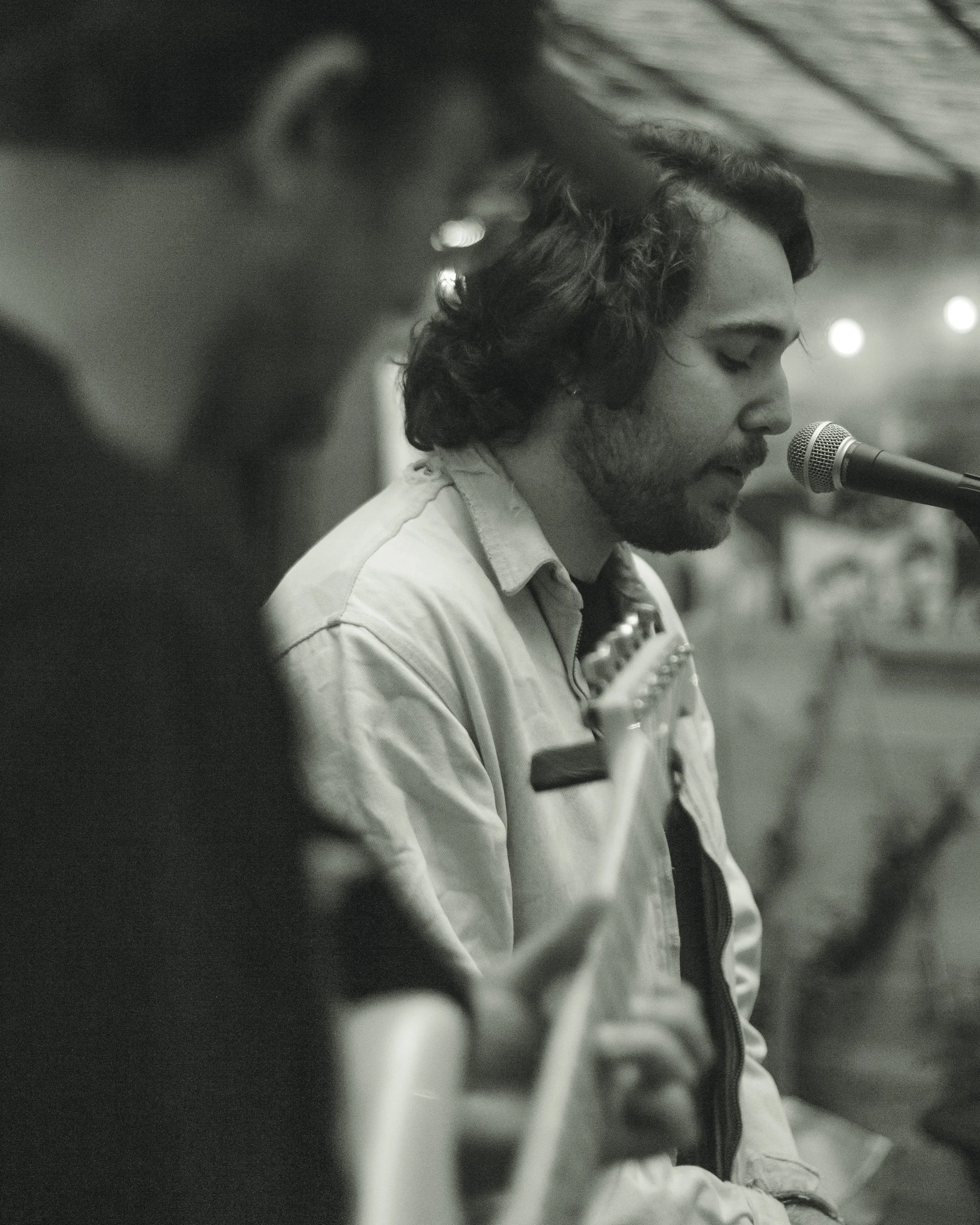 A man with shoulder-length curly hair and a beard is singing into a microphone. He is wearing a light-colored jacket with a zipper and a shirt underneath. The background is blurry, with some lights visible, indicating a possible performance or record