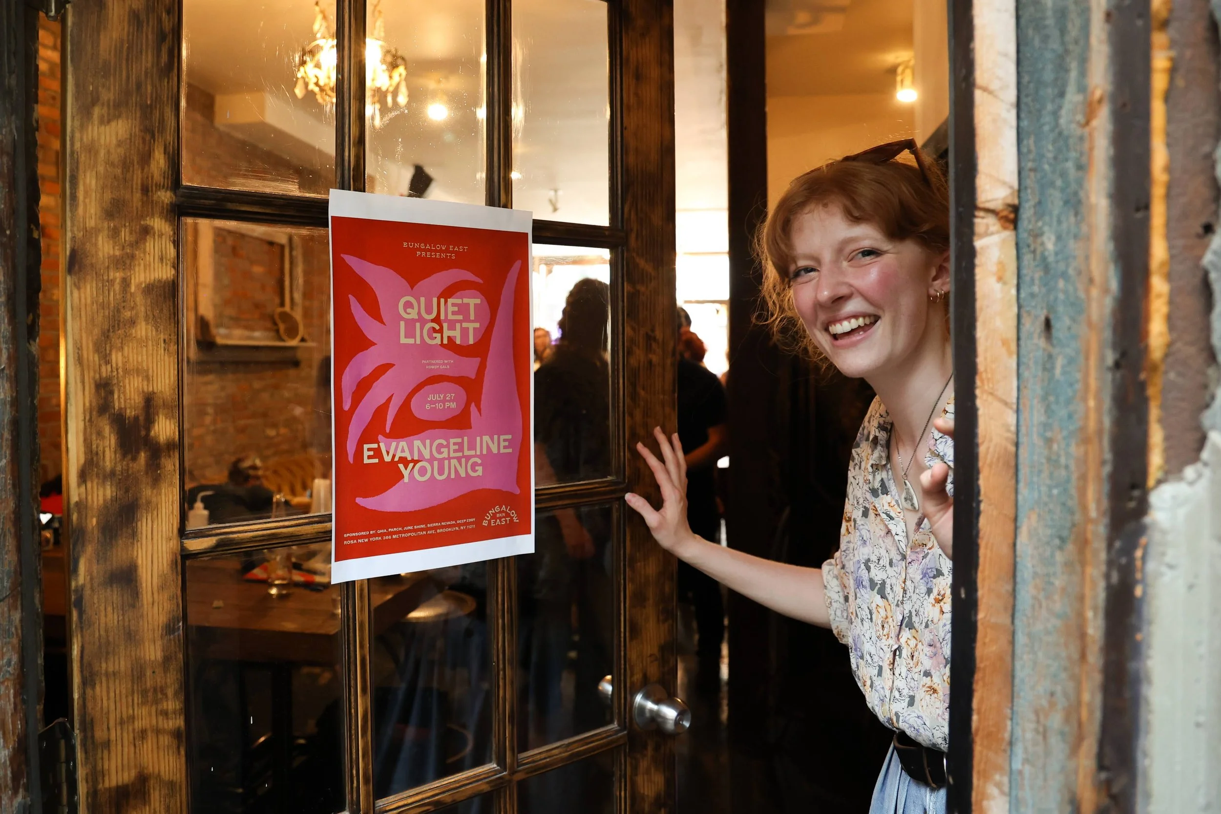 A woman with red hair and earrings peeking through the door of a restaurant or venue with a big smile. There is a poster on the door advertising an event called 'Quiet Light' featuring Evangeline Young, scheduled for July 27 from 6-10 PM, and the wom