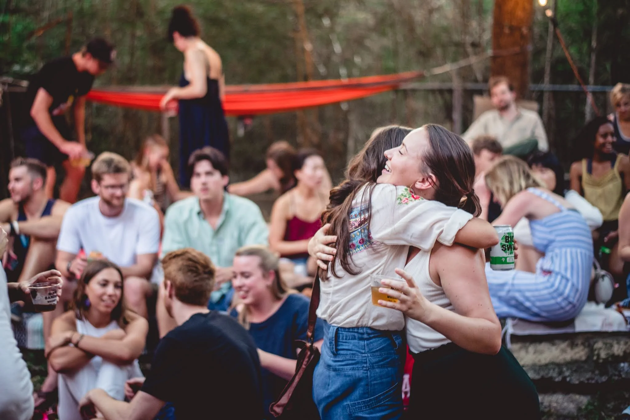 People at an outdoor gathering in a forest, hugging, smiling, and enjoying drinks, with others socializing around them.