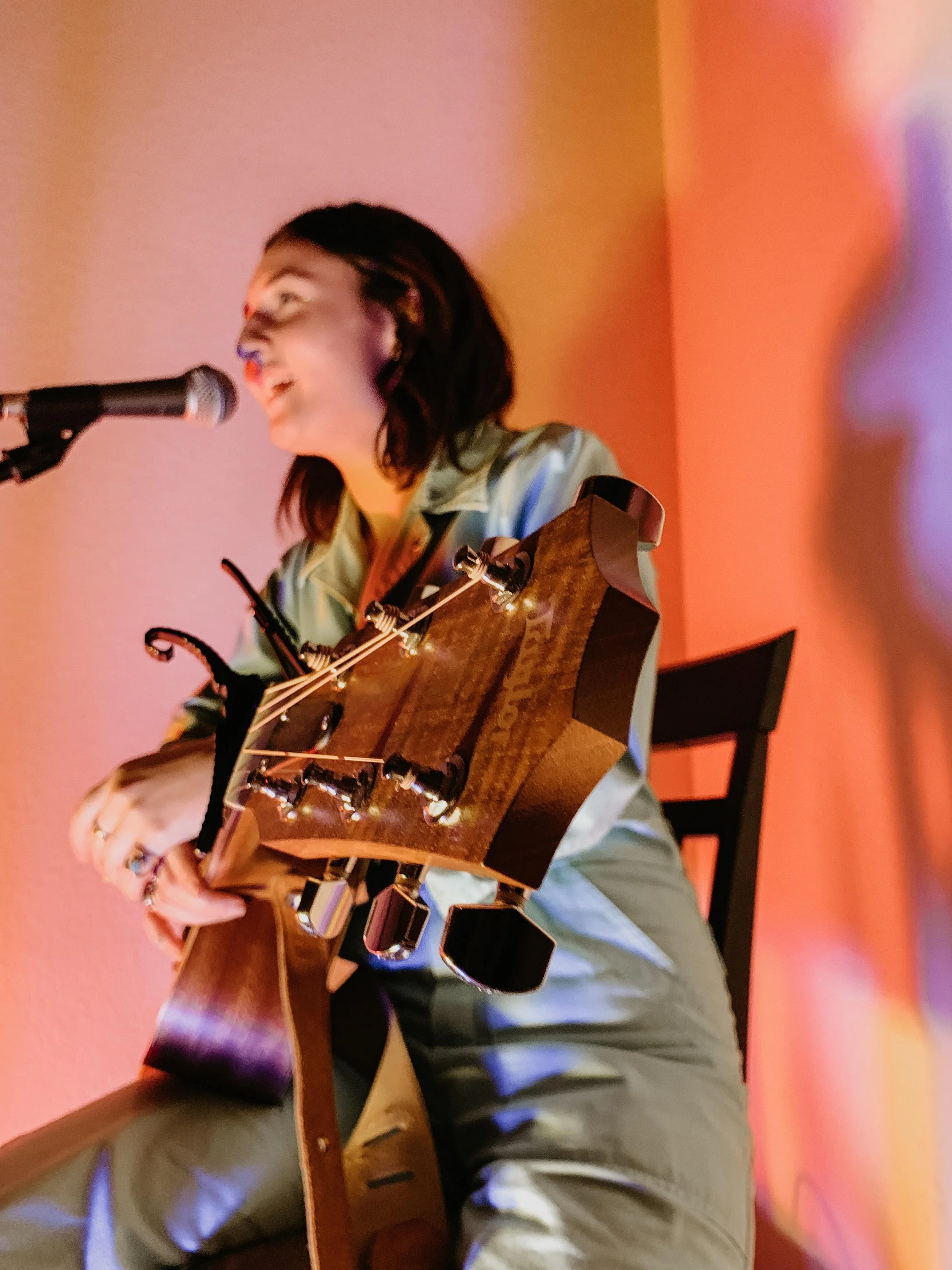 A woman with dark hair singing into a microphone while playing an acoustic guitar, seated on a wooden chair against a colorful background.