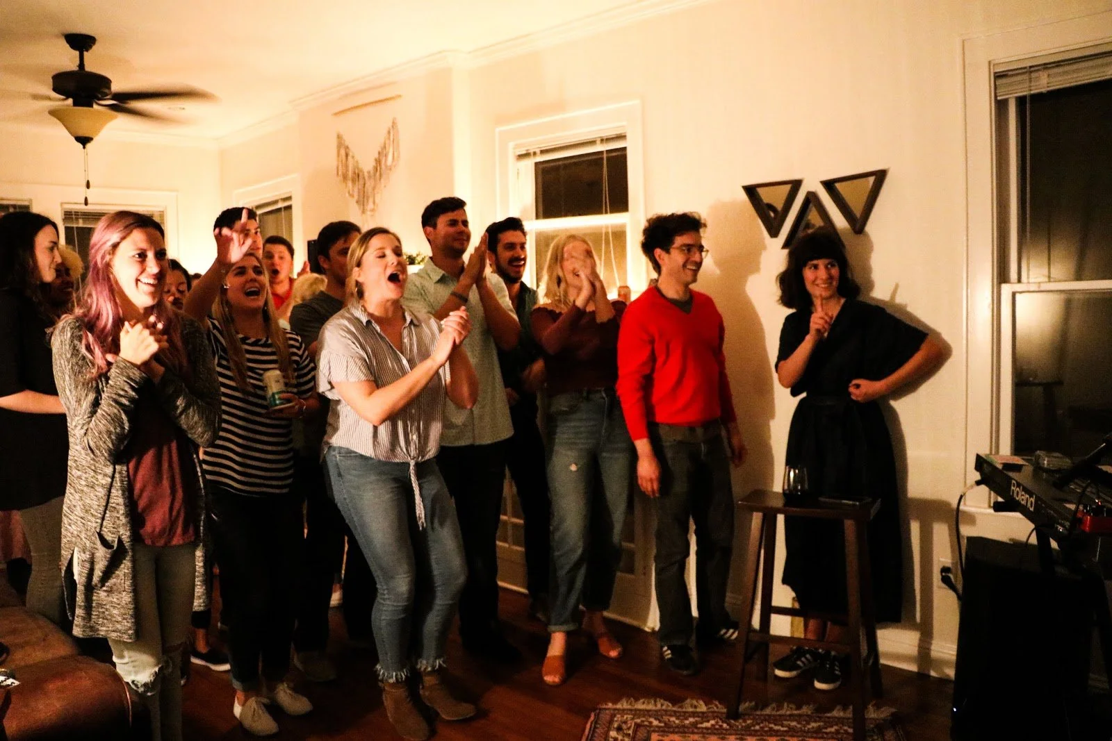 Group of people laughing and smiling in a living room, some with drinks, watching a woman in black standing near a window with geometric wall decorations.
