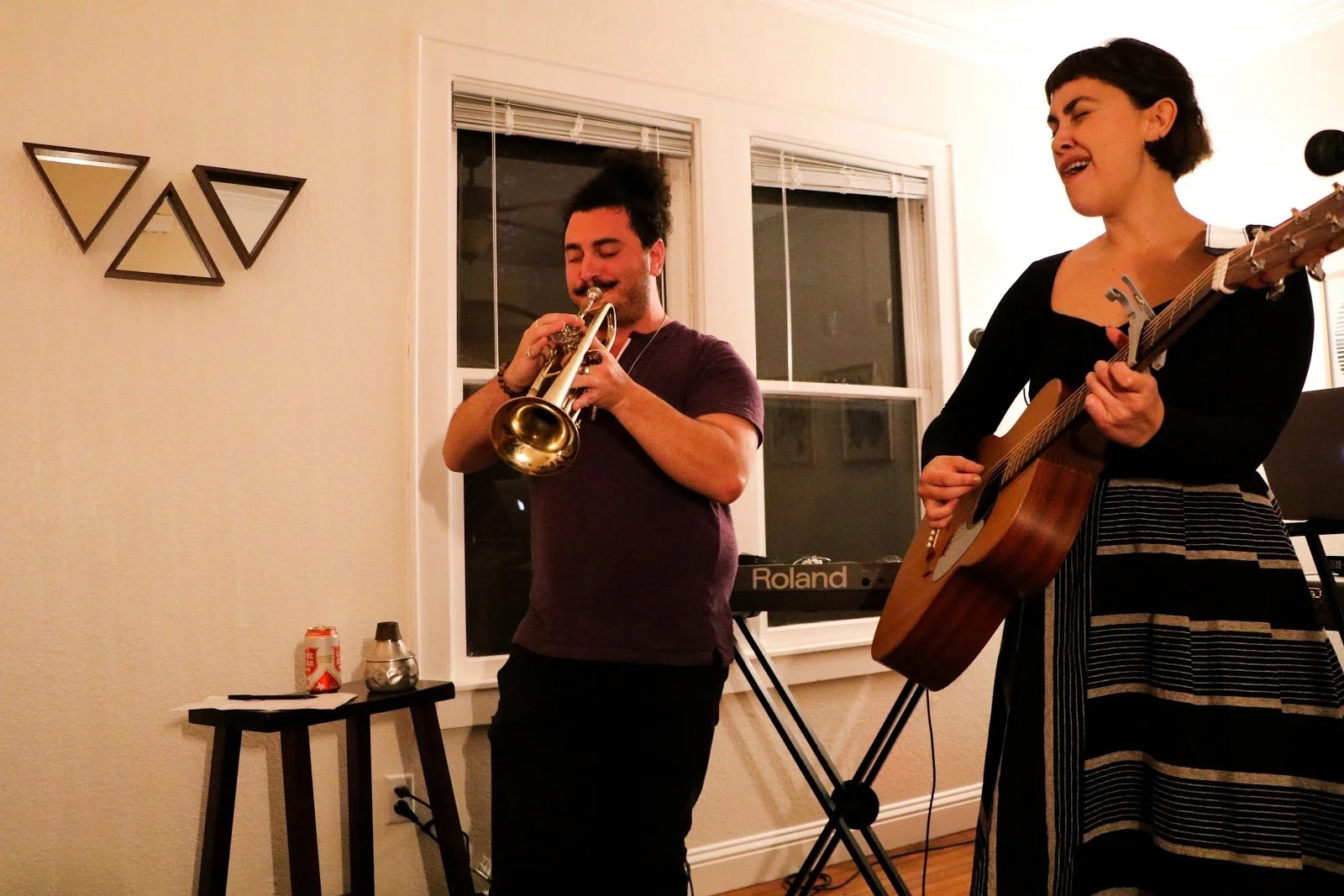Two women playing music indoors, one on trumpet and one on acoustic guitar, with a keyboard in the background and a small table with drink cans and a container next to it.
