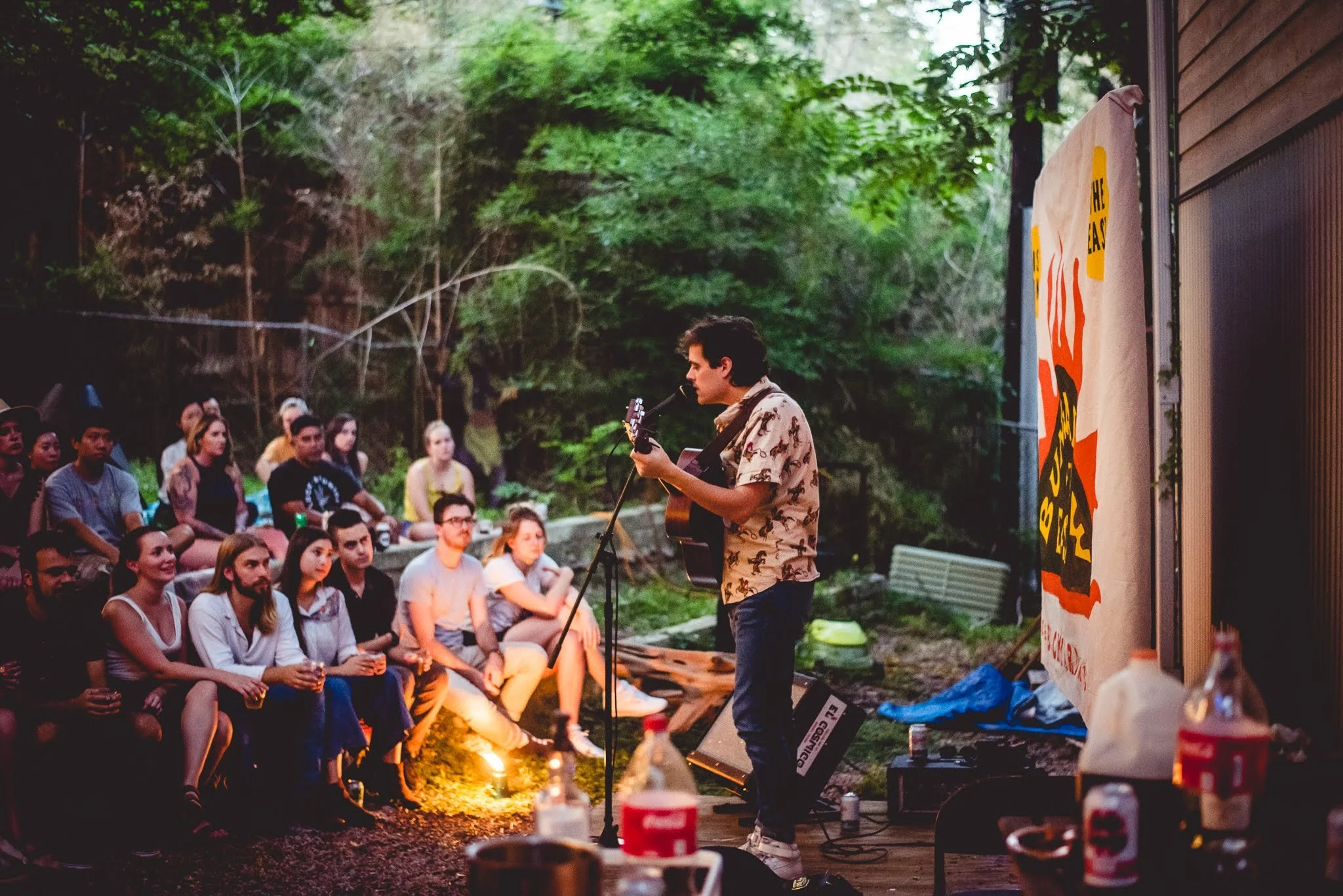 A musician with dark hair playing an acoustic guitar and singing into a microphone outdoors during evening, with an audience sitting on wooden benches surrounded by trees and greenery.