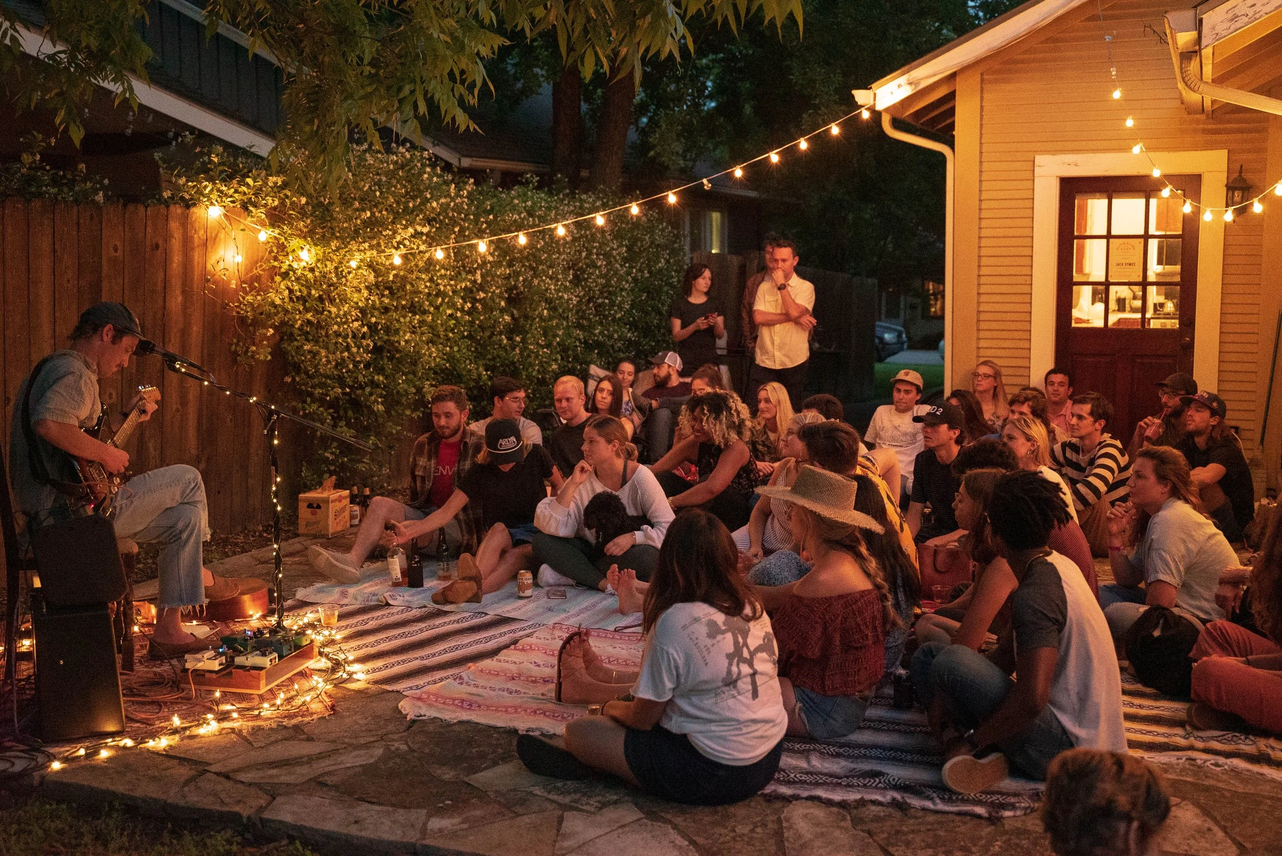A lively outdoor gathering at dusk with a crowd of people sitting on blankets and cushions on the ground, watching a musician playing guitar. String lights are hung above, illuminating the scene, with a yellow house on the right and trees in the back