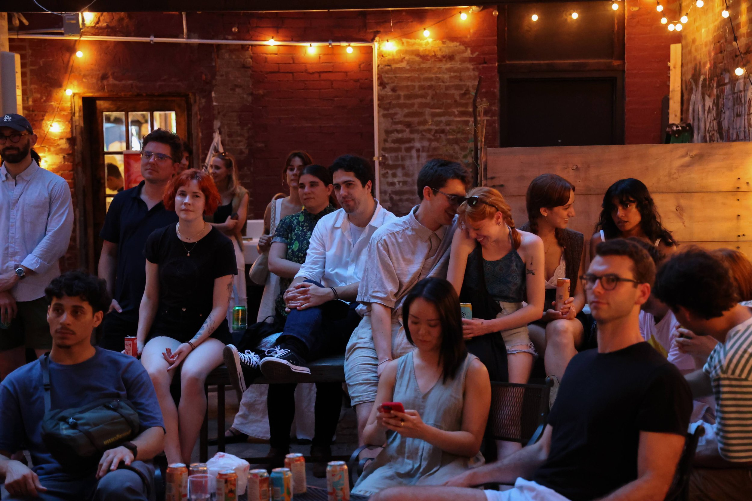 A group of people sitting and standing in a cozy, dimly lit venue with exposed brick walls and string lights, attending a social event or gathering.