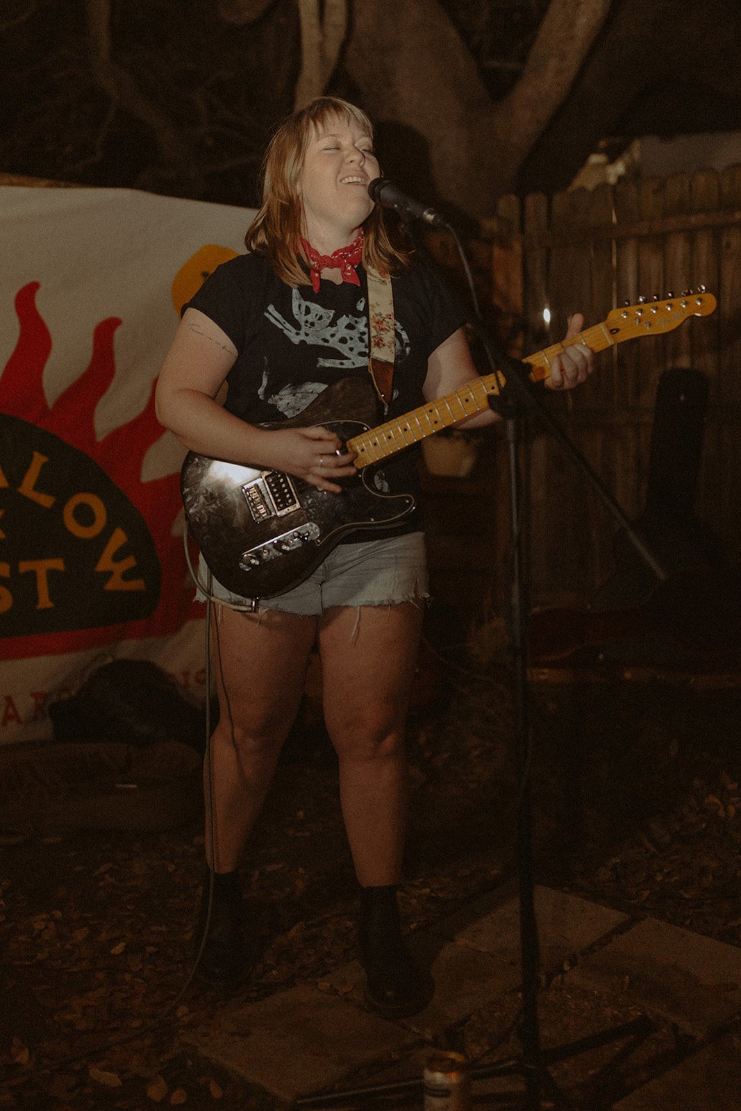 A woman with red hair singing and playing an electric guitar on stage, in a dimly lit setting with a wooden wall and a banner with flames in the background.