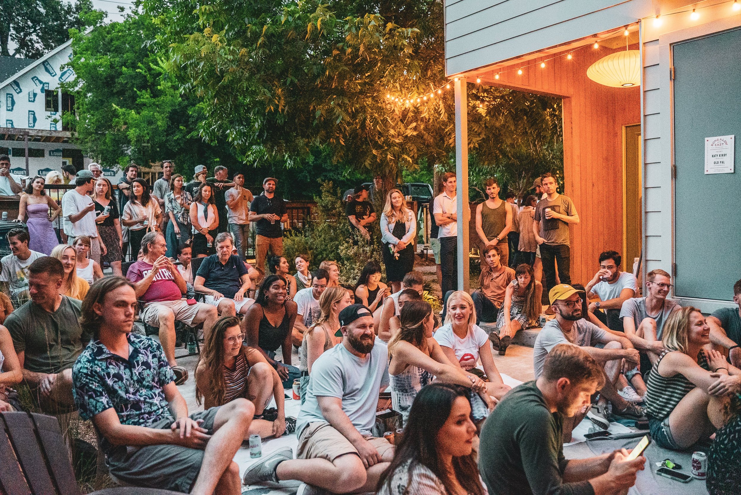 A crowd of people gathered in an outdoor backyard setting, sitting and standing, watching a show or event, with string lights and a wooden structure with hanging lanterns.