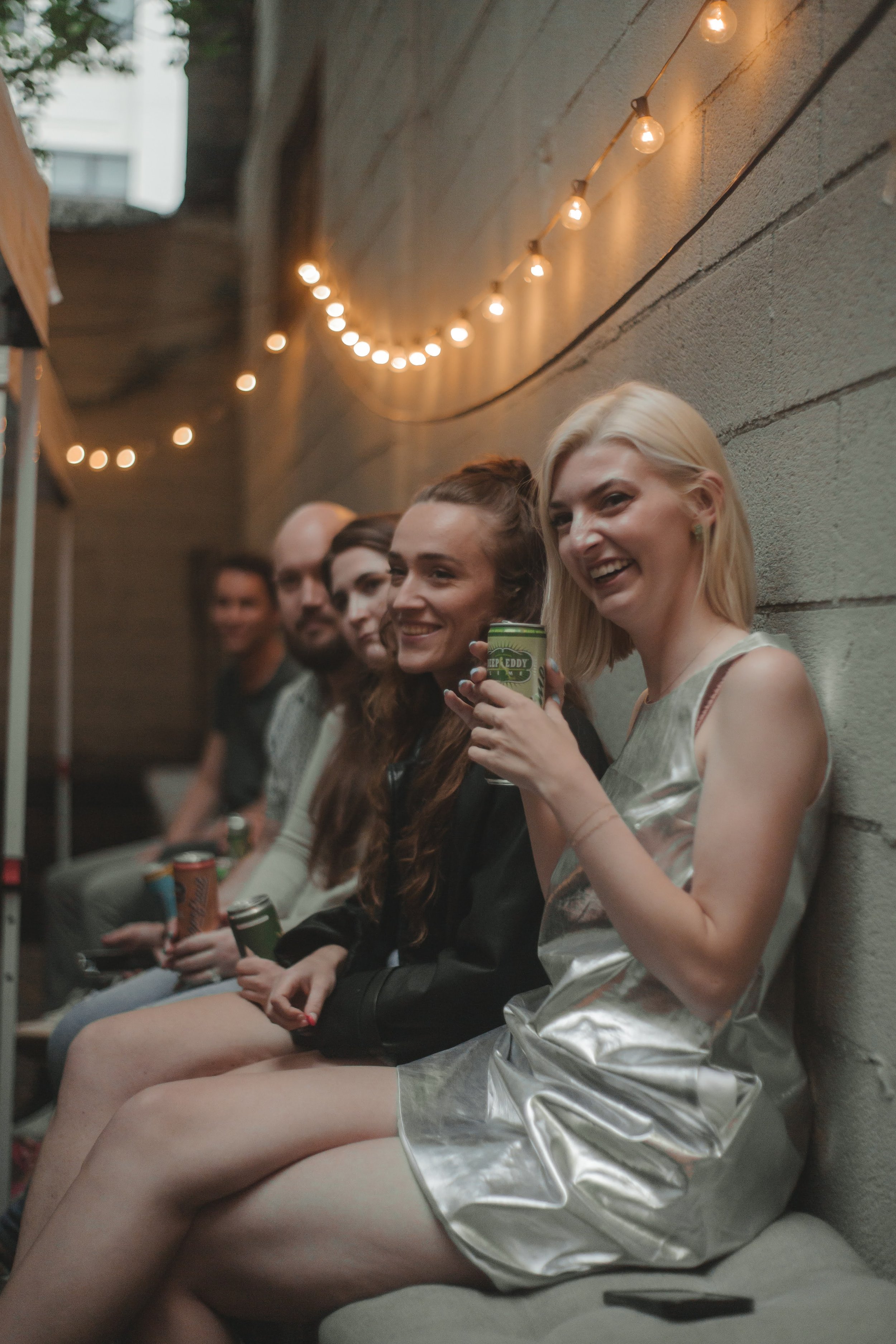 Group of people sitting on a bench against a concrete wall, smiling, with string lights hanging above them and holding drinks.