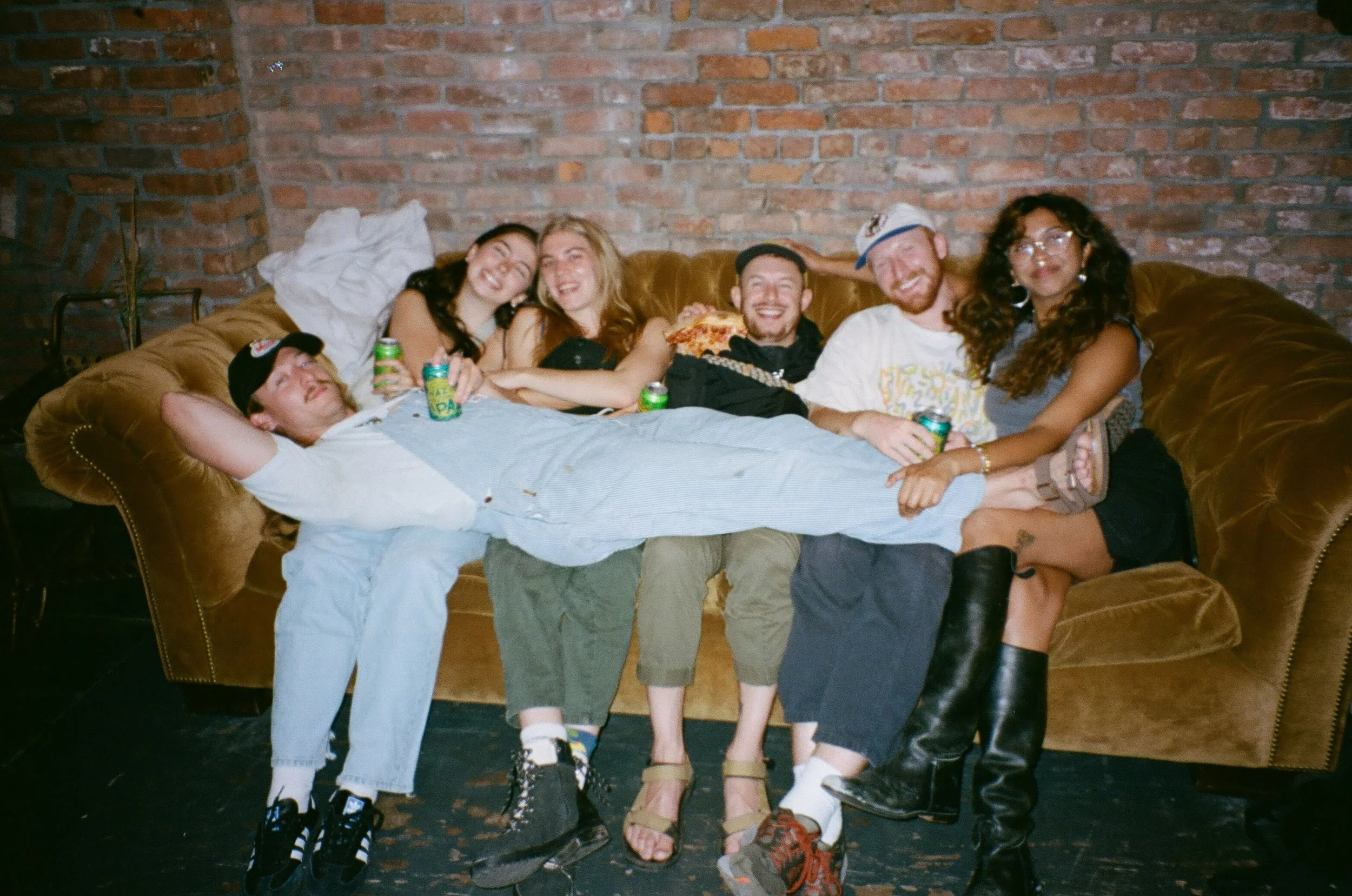Group of six friends sitting on a brown leather couch against a brick wall, smiling and holding drinks and pizza slices.