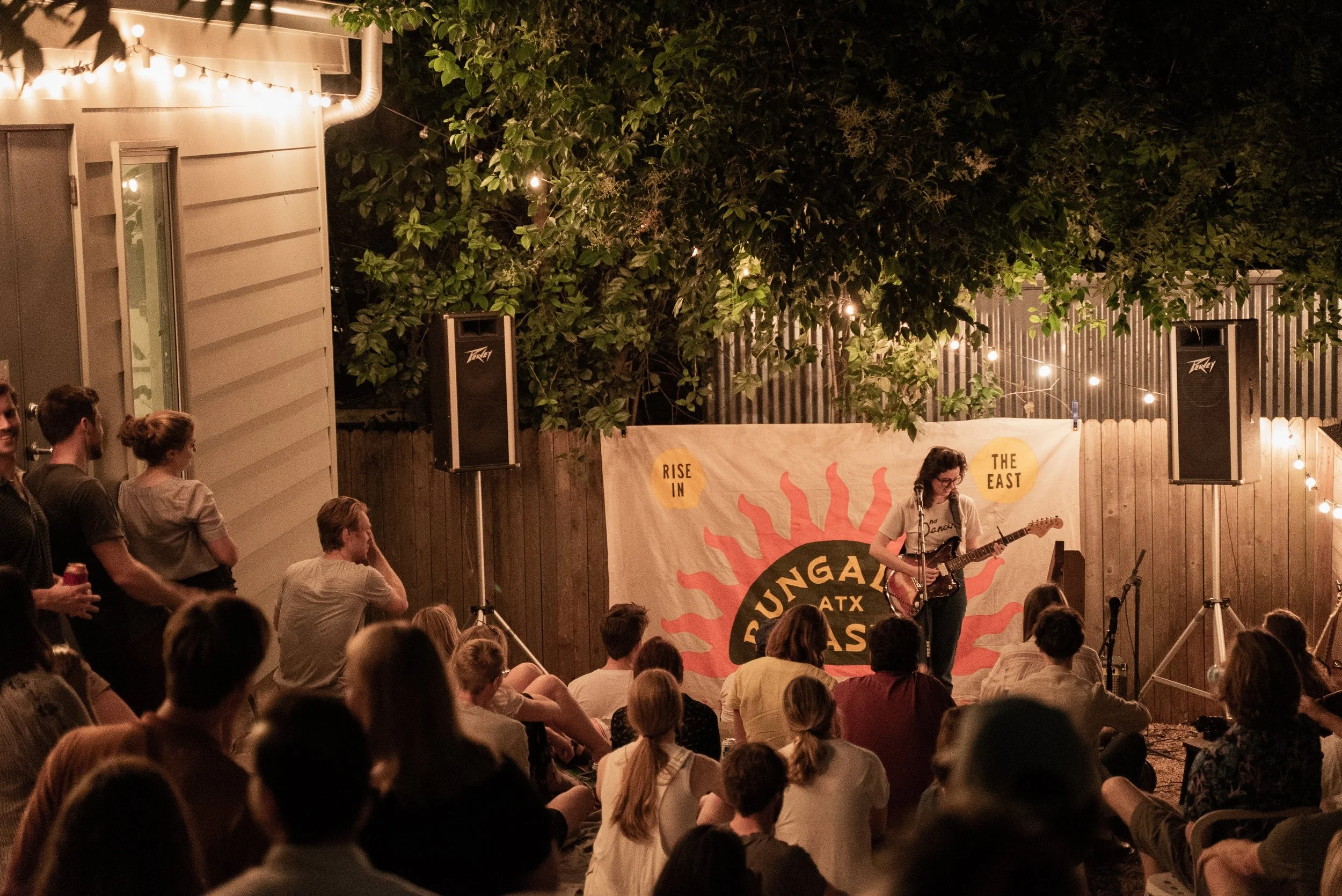 A nighttime outdoor concert with a crowd gathered in front of a small stage. A musician is playing an electric guitar, and there are string lights above and trees in the background. The setting appears to be a backyard party or similar event.