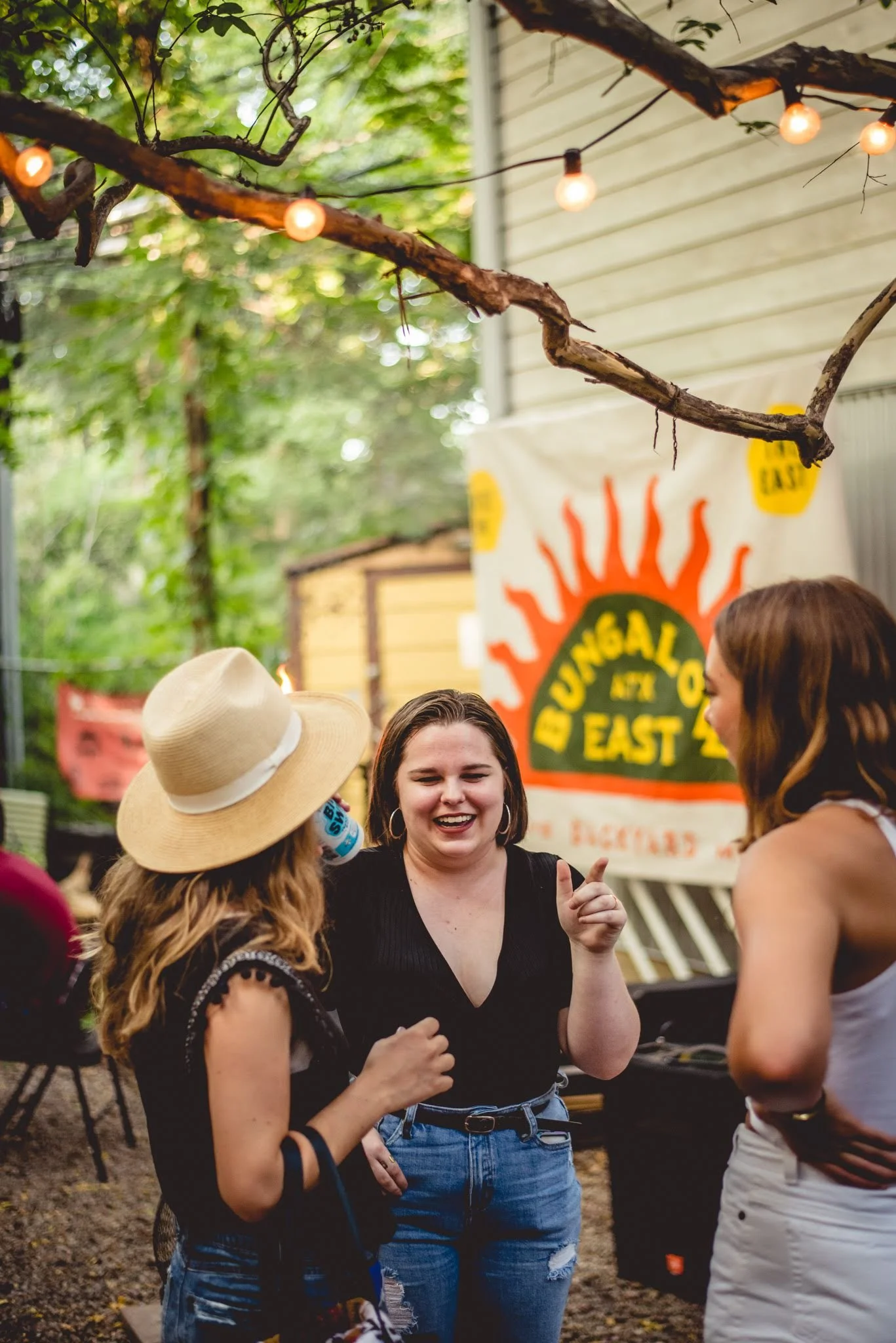 Four women are engaged in conversation outdoors, with a large colorful sign in the background indicating a festival or event. One woman is wearing a straw hat and has long, curly hair. Another woman is smiling, wearing a black top and high-waisted je