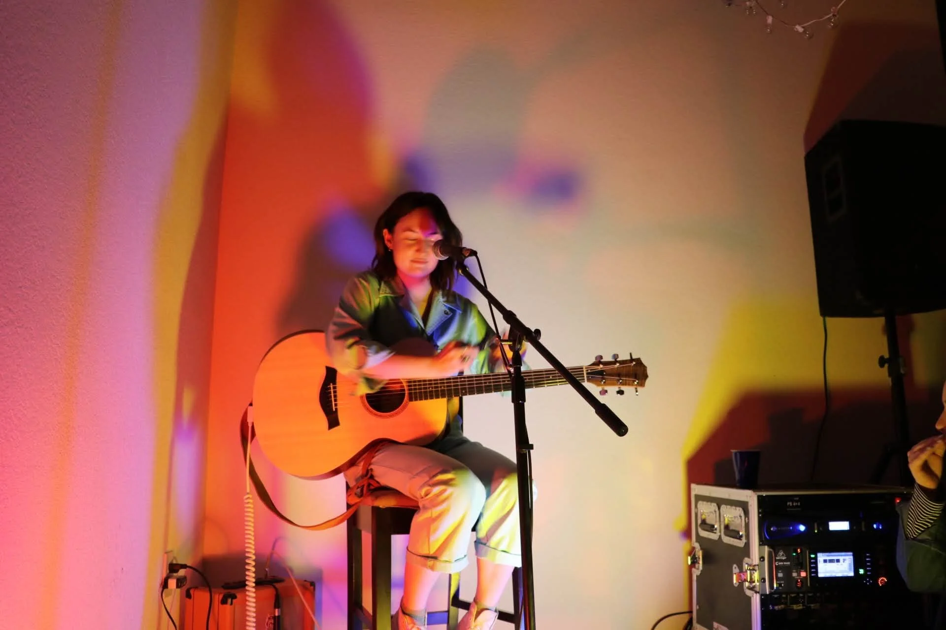 A woman sitting on a stool playing an acoustic guitar and singing into a microphone on a small stage, with colorful stage lighting and musical equipment visible.