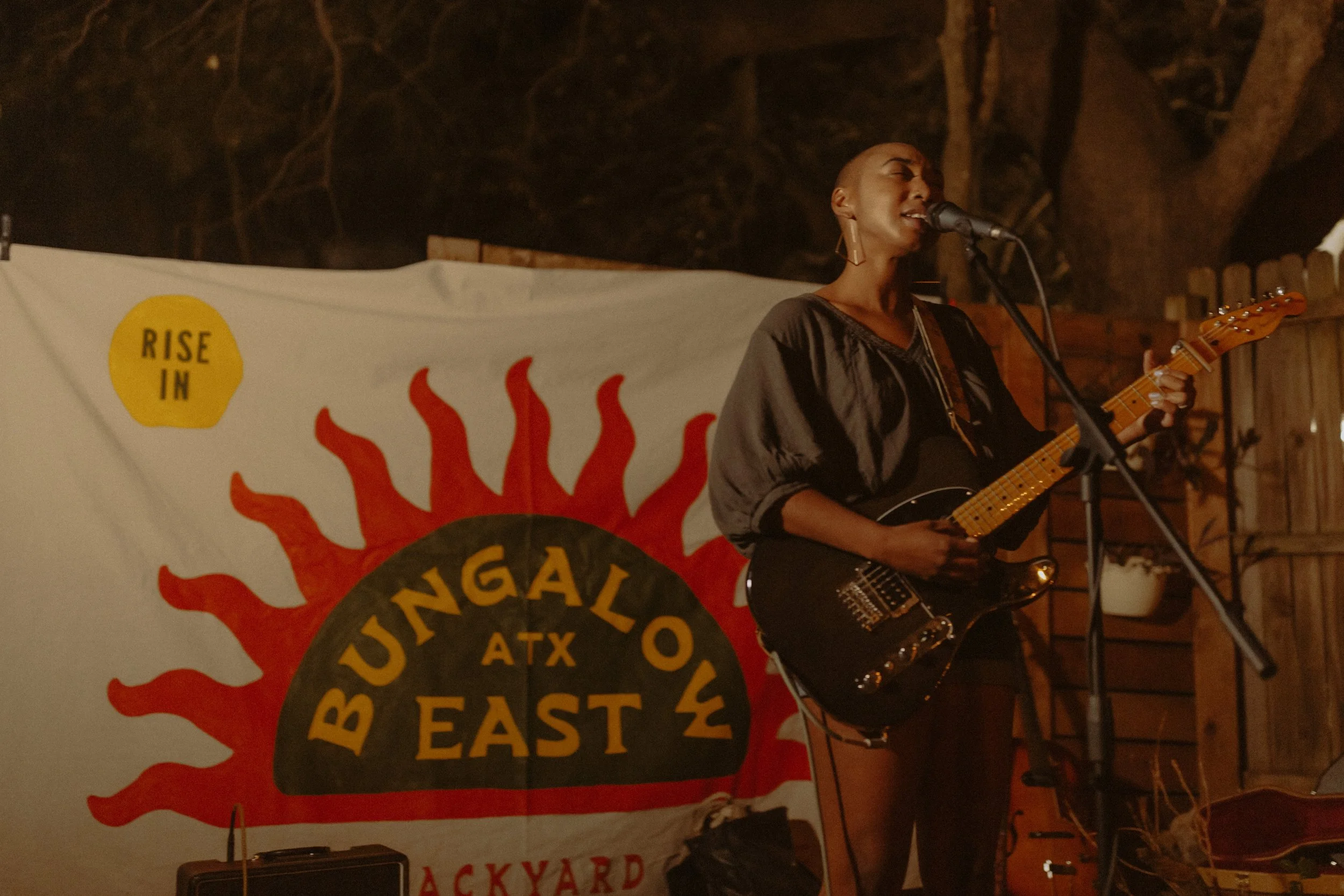 A woman singing and playing guitar on stage at a music event, with a banner that reads 'Rise in' and 'Texas Beach' in the background.