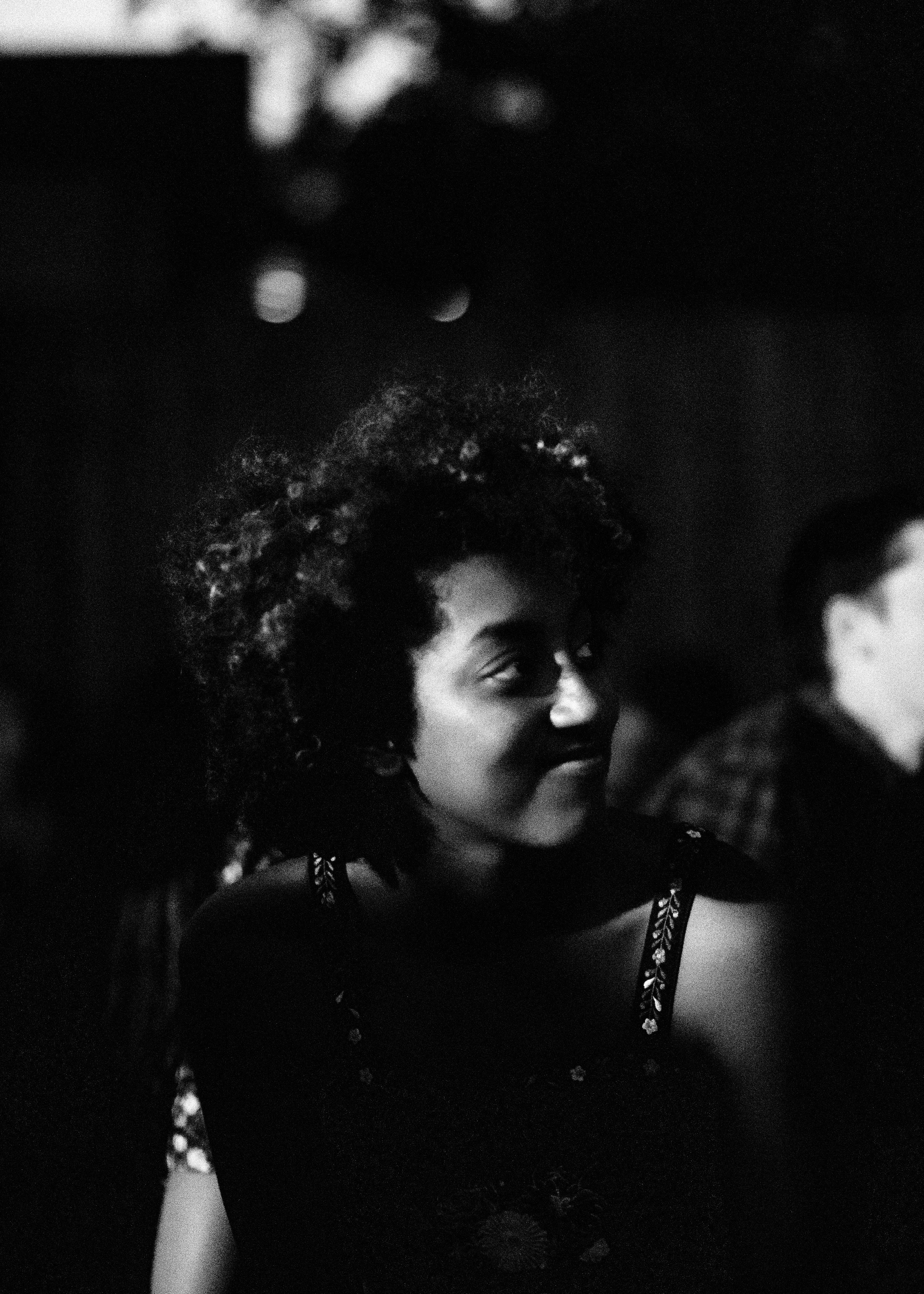 A woman with curly hair smiling in a dark setting, wearing a dress with patterned straps.