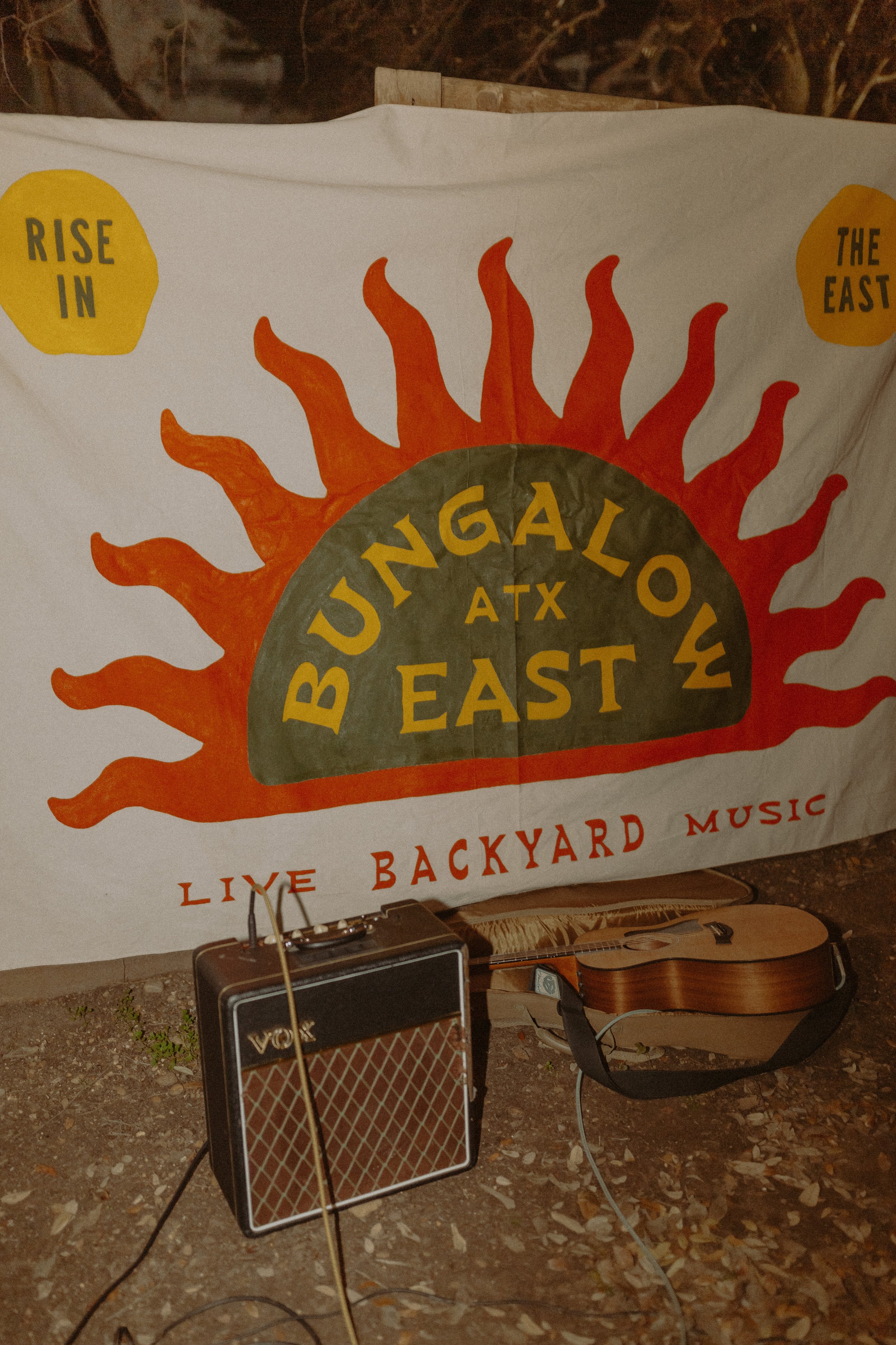 A flag with orange and yellow text reading "Bungalow East" with a sunburst graphic, a Vox guitar amplifier, and an acoustic guitar on the ground among leaves, set outdoors at night.