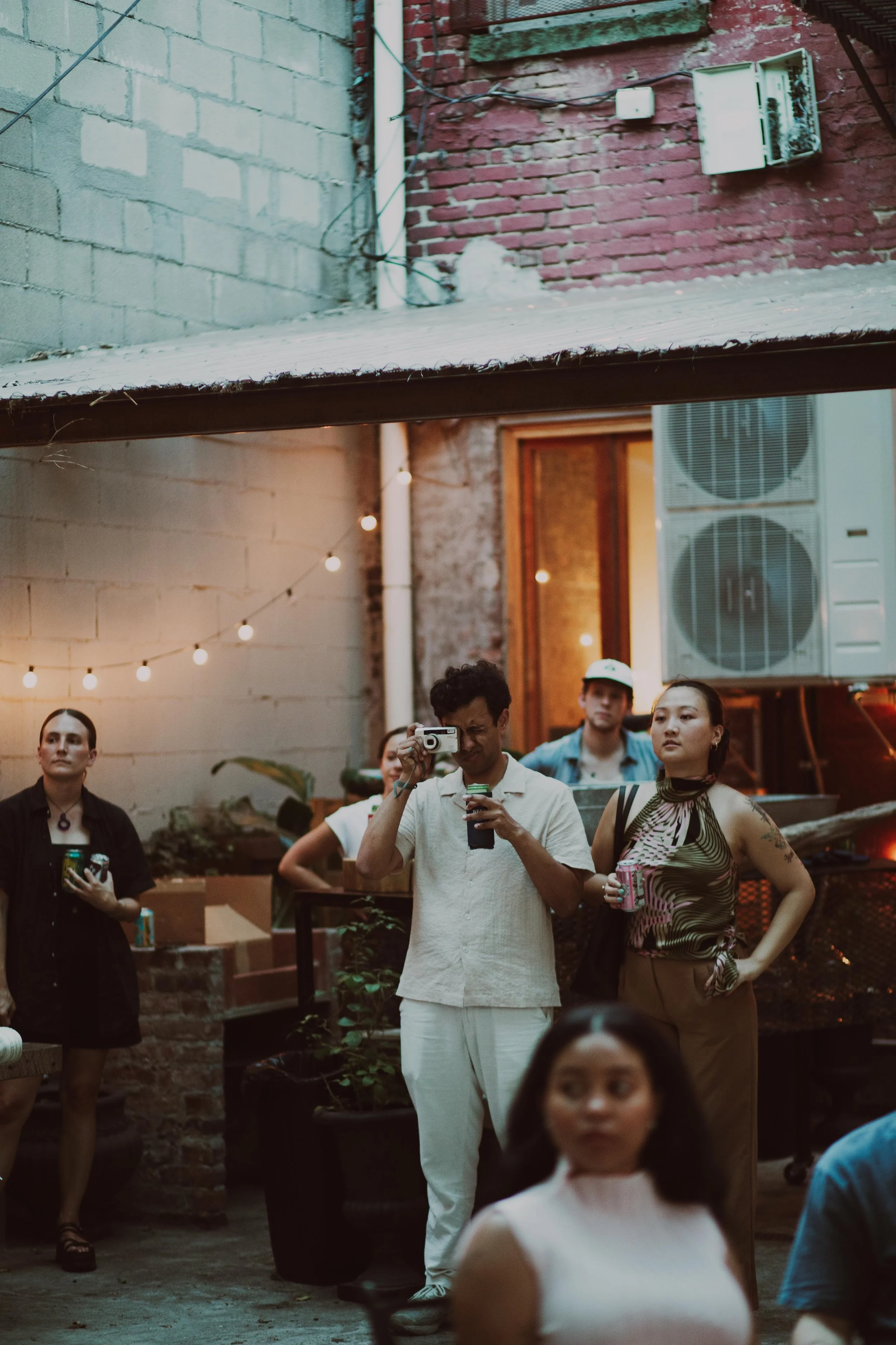 People gathered at an outdoor event in an urban alley with string lights, brick and concrete walls, and air conditioning units visible in the background.