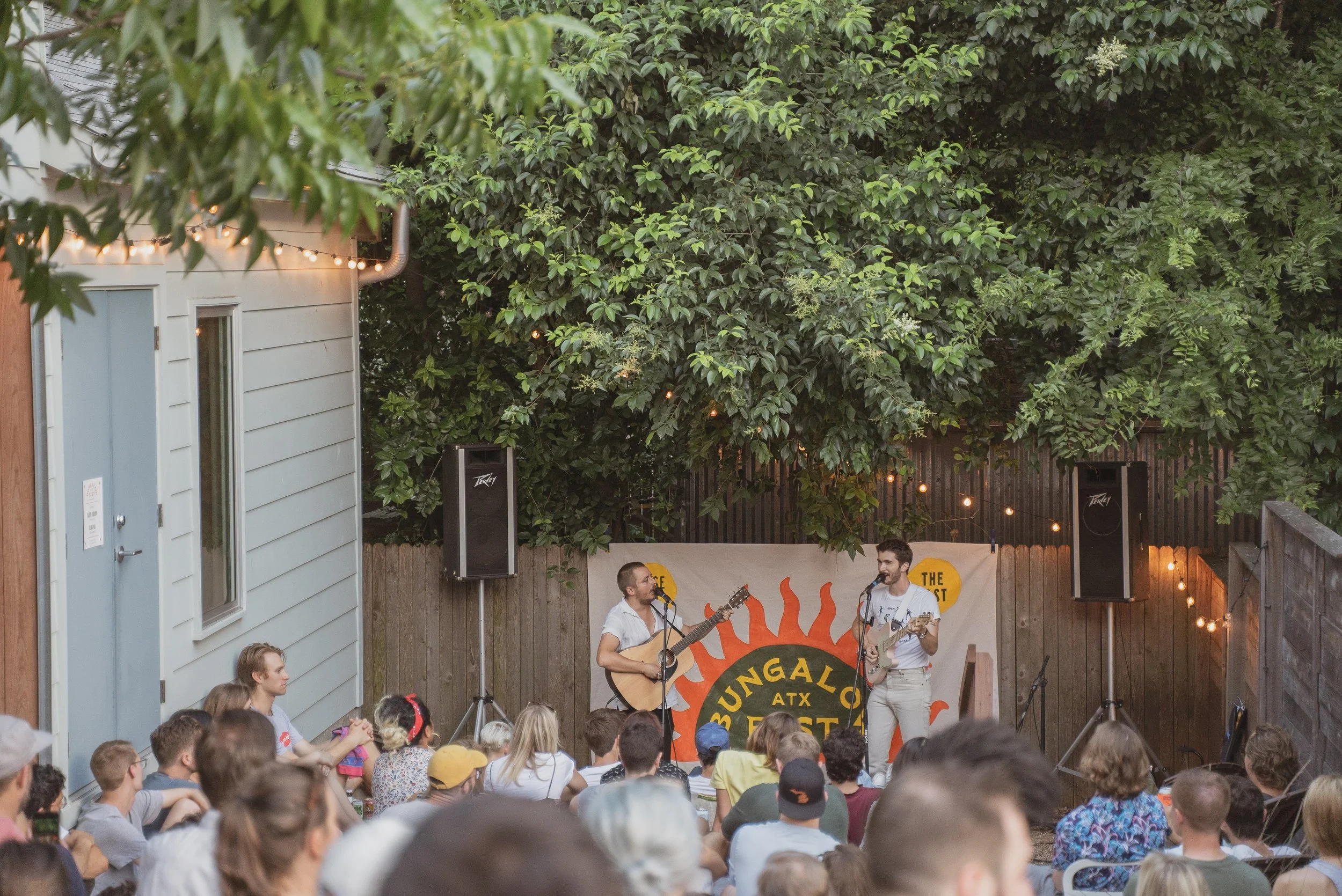 Two musicians performing with guitars on a small outdoor stage surrounded by an audience in a backyard. There are string lights and a banner in the background.