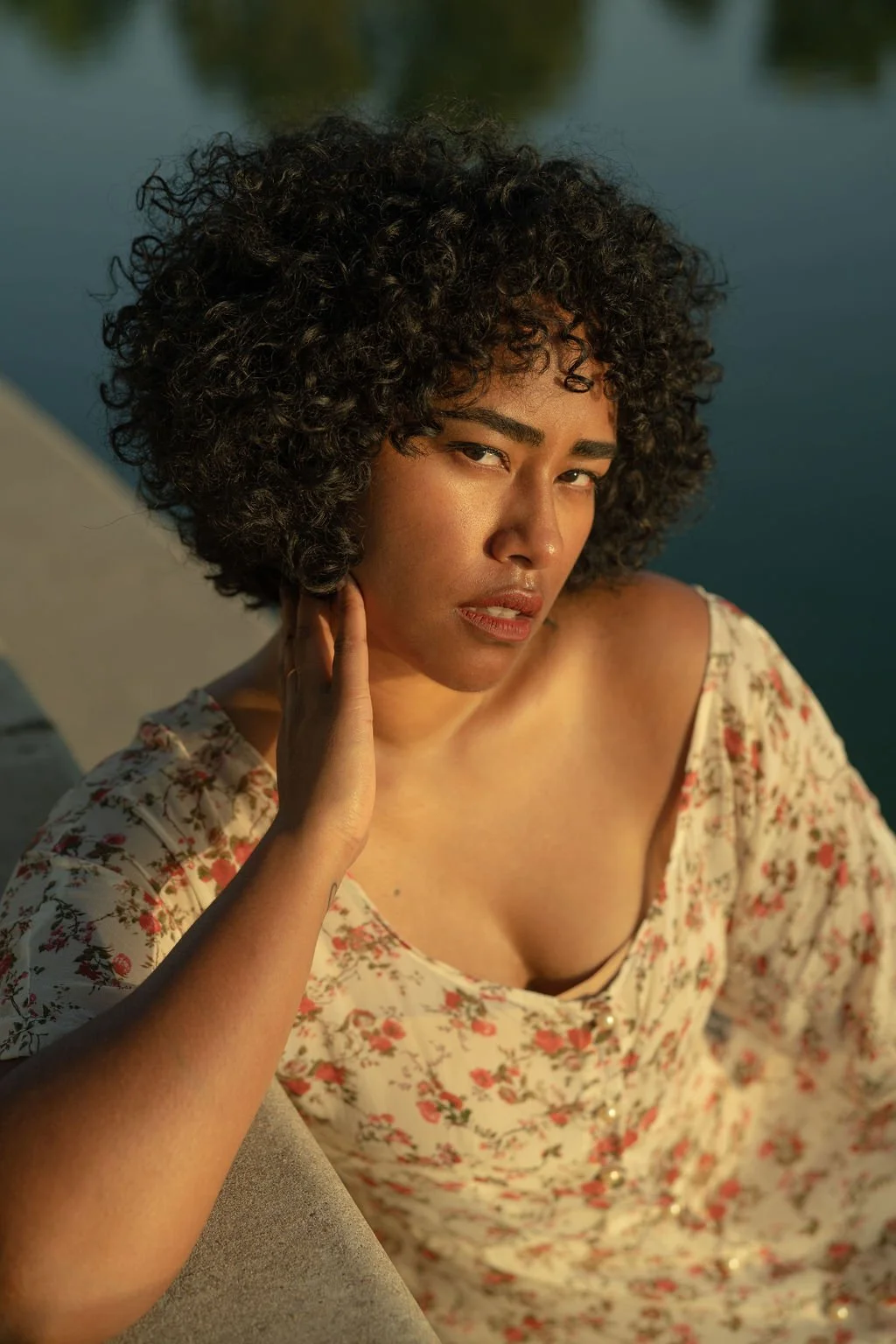 A woman with curly black hair, wearing a floral dress, sitting near water, looking directly at the camera.