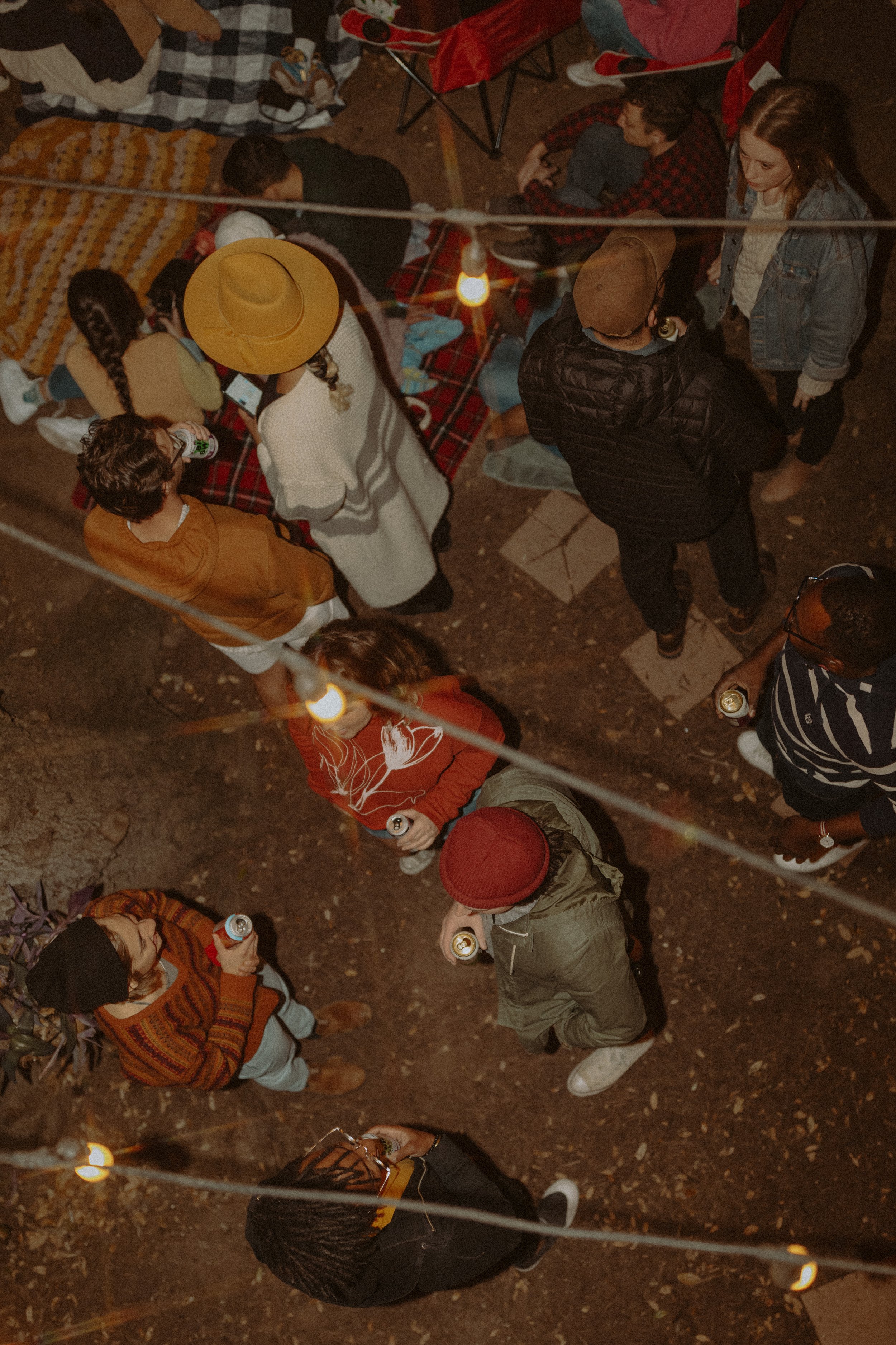 People gather outdoors at night, some sitting on blankets, socializing and holding drinks. There are string lights overhead, creating a cozy atmosphere.