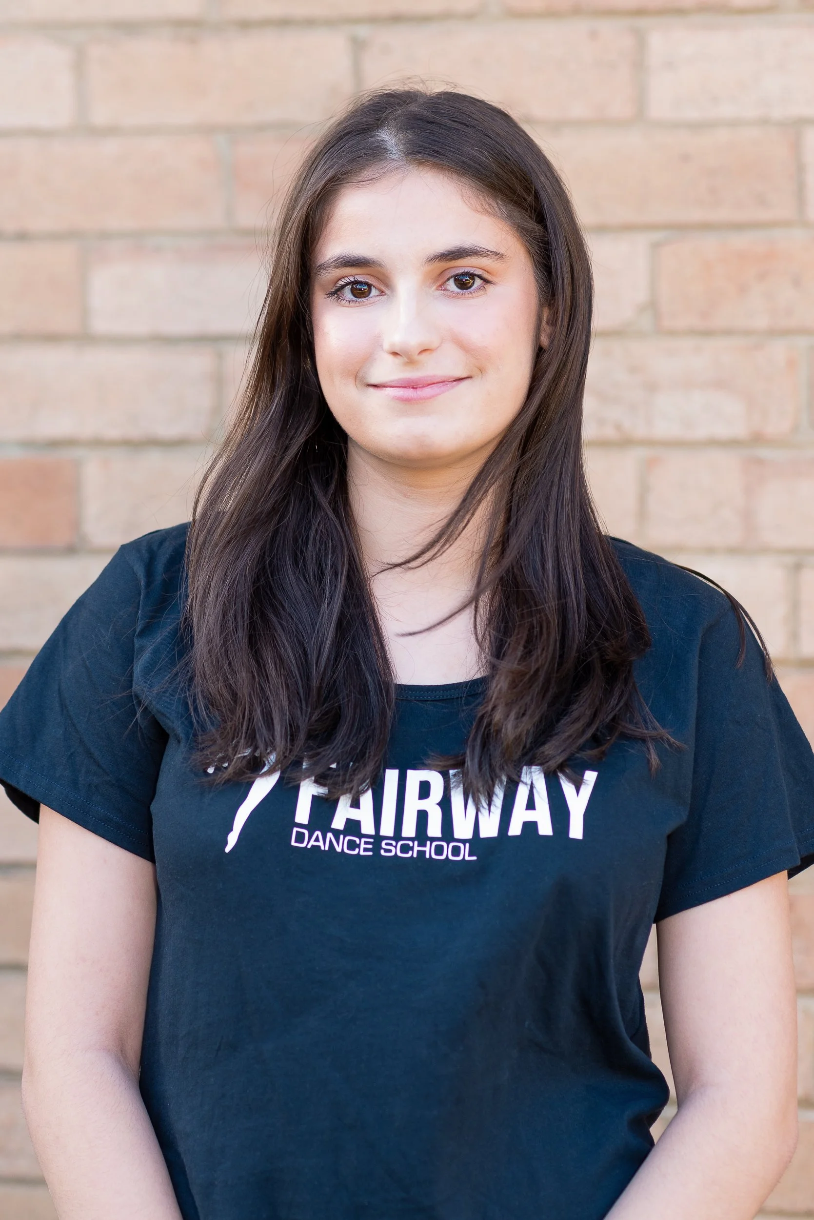 Smiling young woman with long dark hair wearing a black t-shirt with 'Fairway Dance School' logo, standing in front of a brick wall.