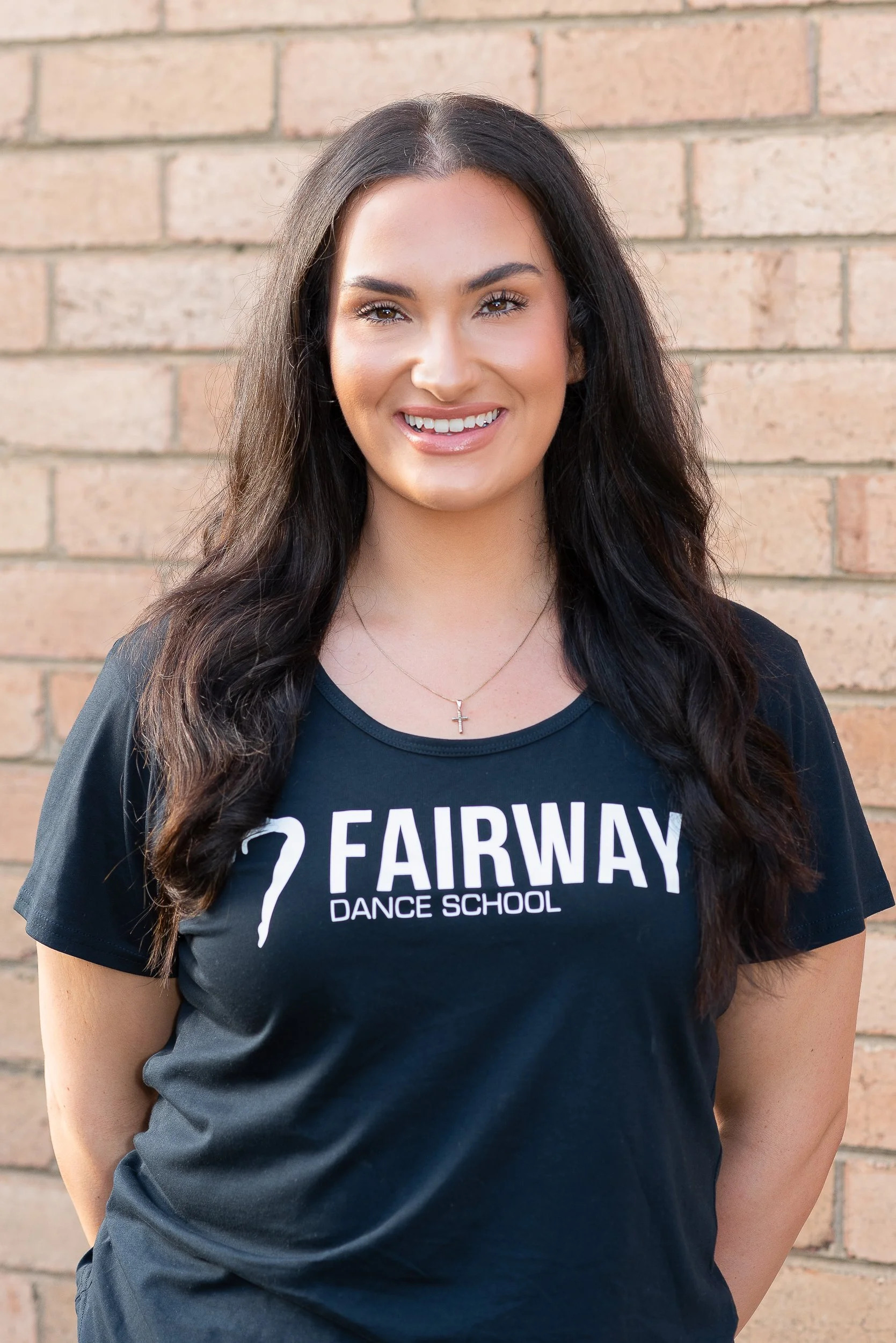 Young woman with long dark hair smiling, wearing a black t-shirt that says 'Fairway Dance School,' standing against a brick wall.