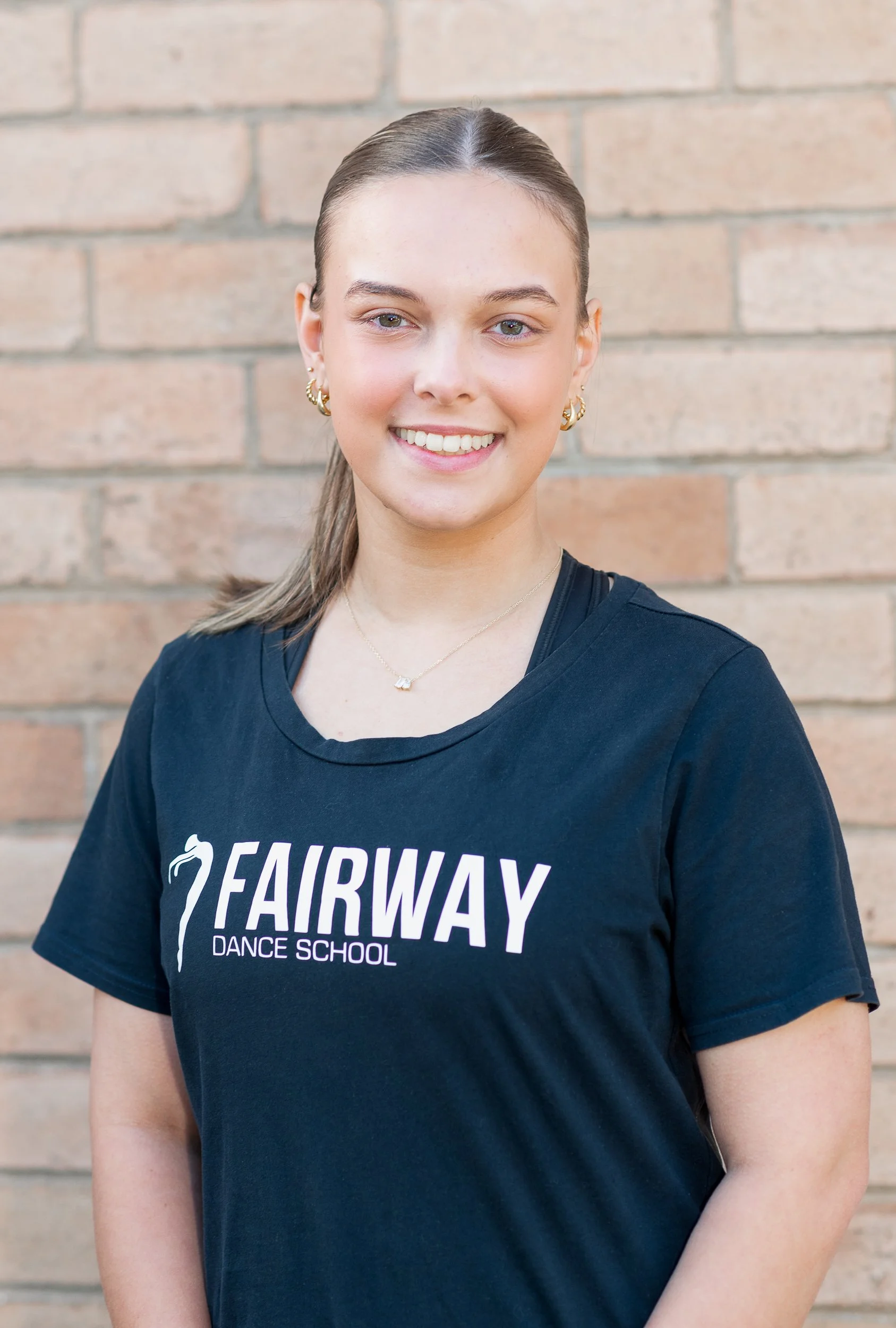 A young woman with light skin, blonde hair tied back, smiling, wearing gold hoop earrings, a necklace, and a black T-shirt that says "FAIRWAY DANCE SCHOOL" with a dance-related logo, standing in front of a brick wall.