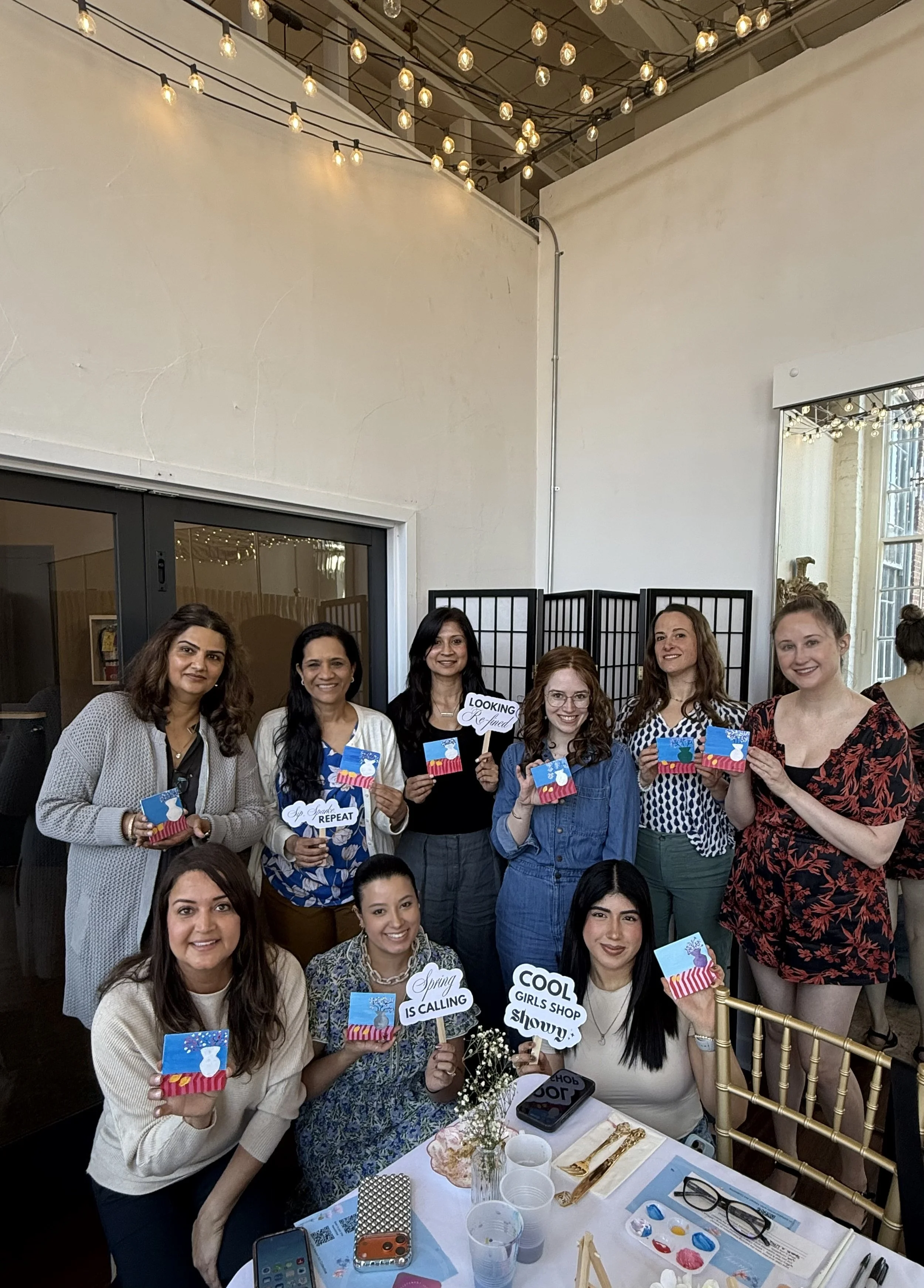Group of women at a celebration, holding colorful gift boxes and signs with cheerful messages, in a decorated indoor setting with string lights.