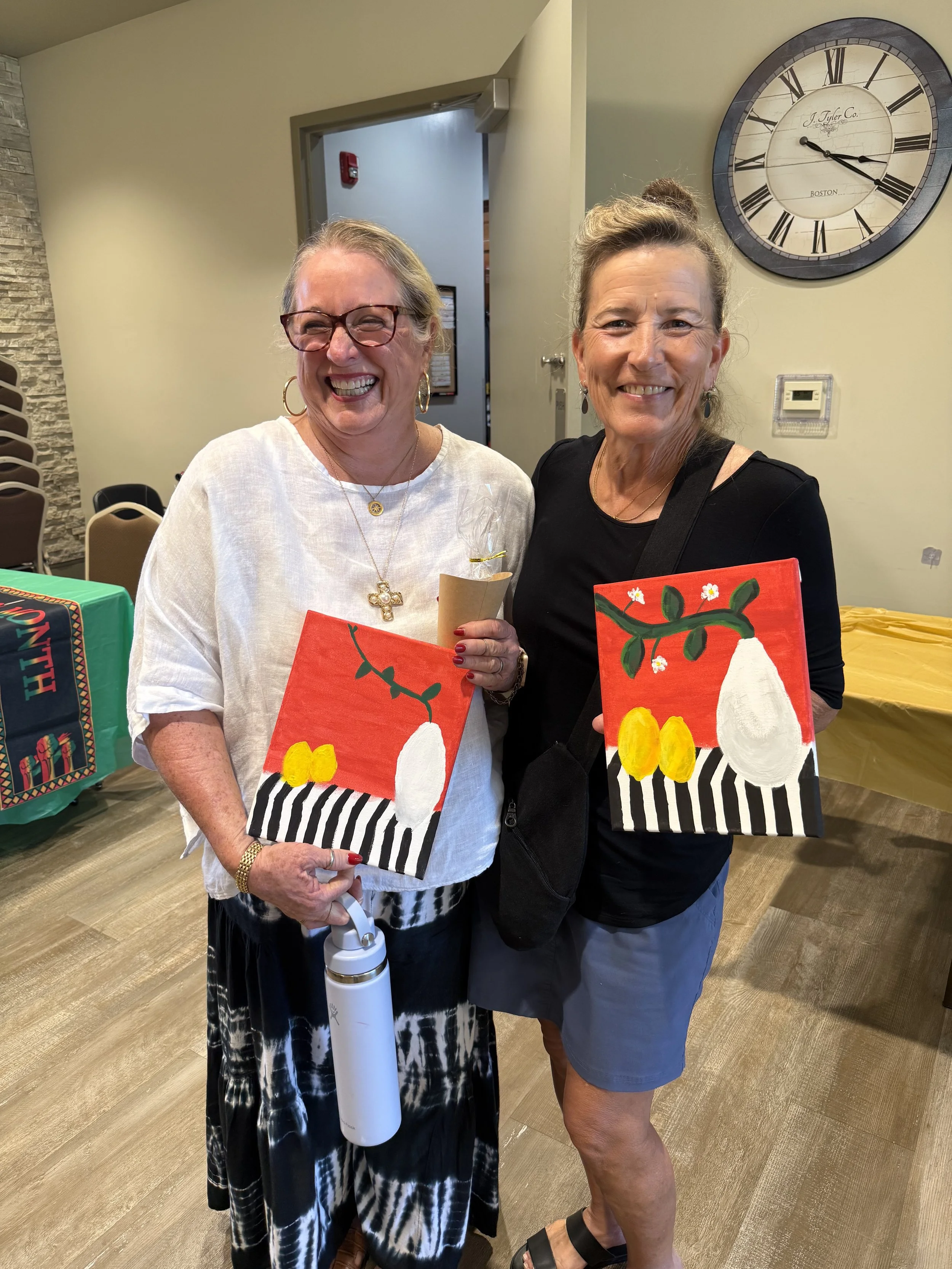 Two smiling women standing indoors, each holding a small painting with a red background, white vase, yellow and white flowers, and green leaves, in front of a wall clock and beige walls.