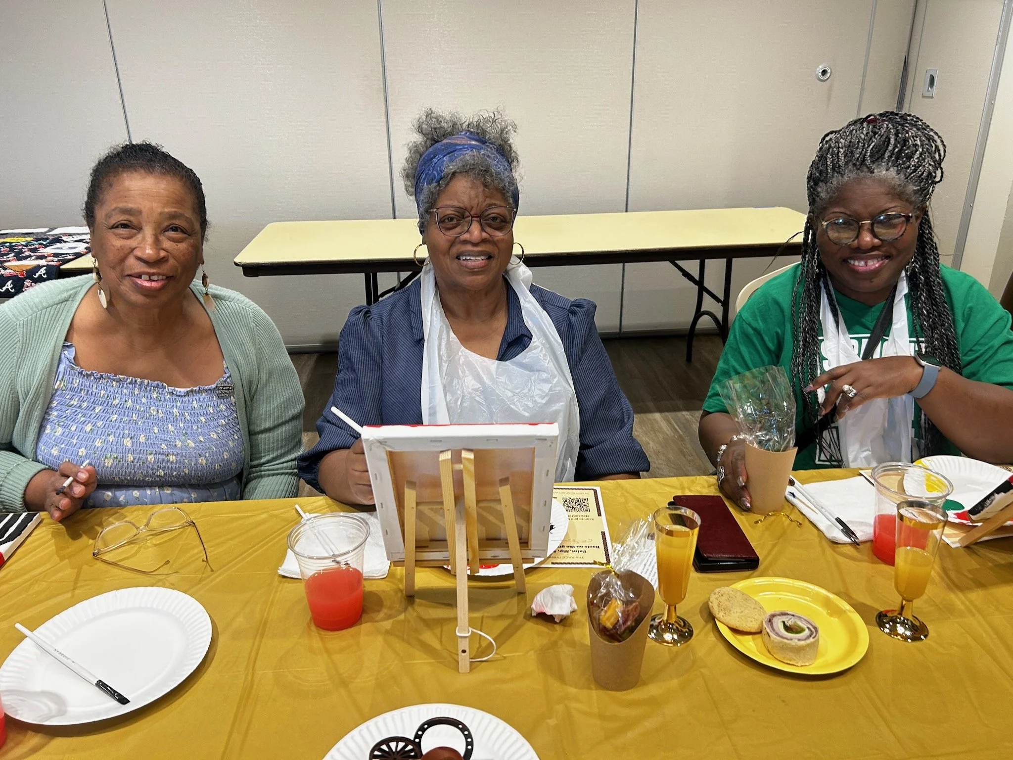Three women sitting at a table during a celebration or gathering, with food and drinks, smiling at the camera.
