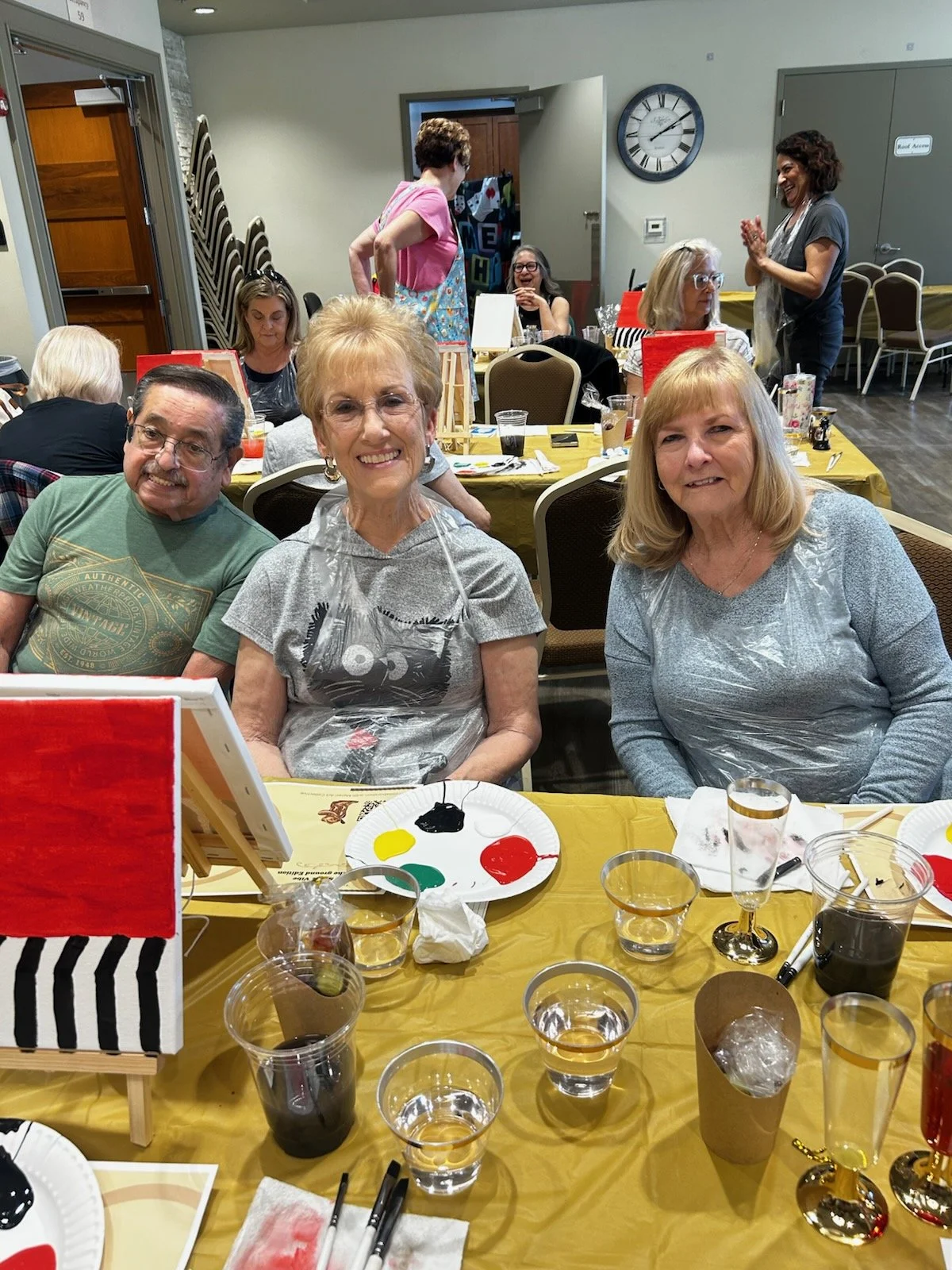 Three older adults sitting at a decorated table during a social gathering or art event, with paintings and art supplies visible on the table, and other guests in the background.