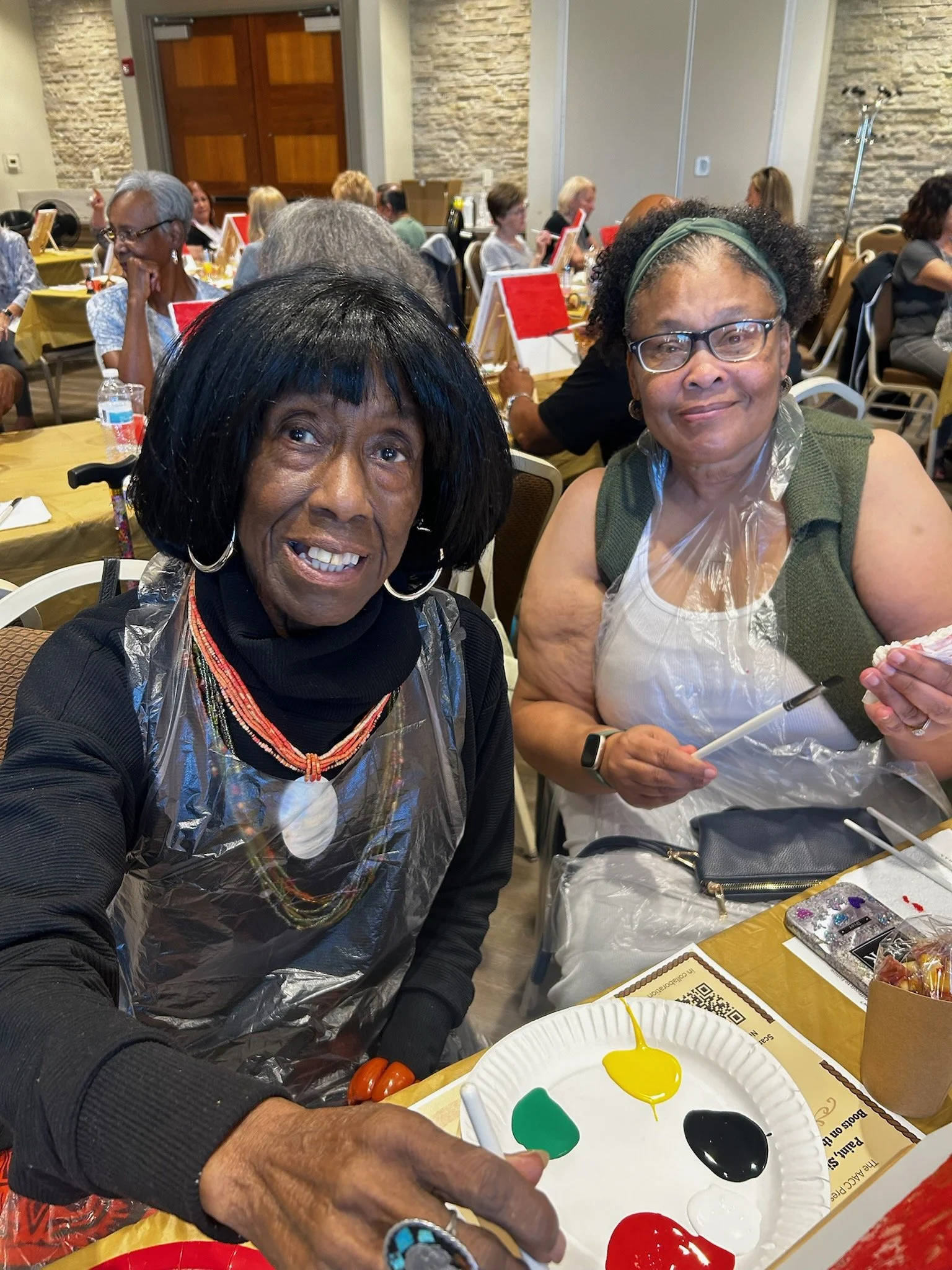 Two women sitting at a decorated table during a group event, smiling at the camera. One woman is wearing a black top, and the other is wearing glasses, a green vest, and a plastic apron. There are multiple people in the background, some looking at ca