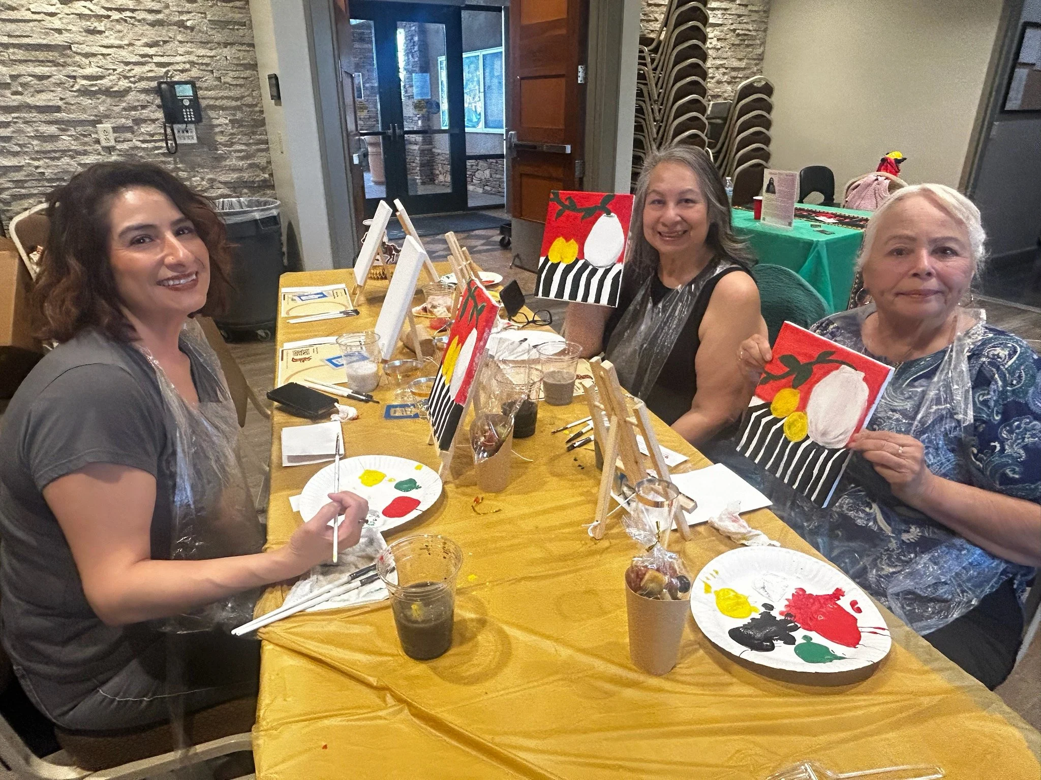 Four women participating in a painting activity at a table, holding up their finished paintings of abstract art with bright colors, in a decorated indoor setting.