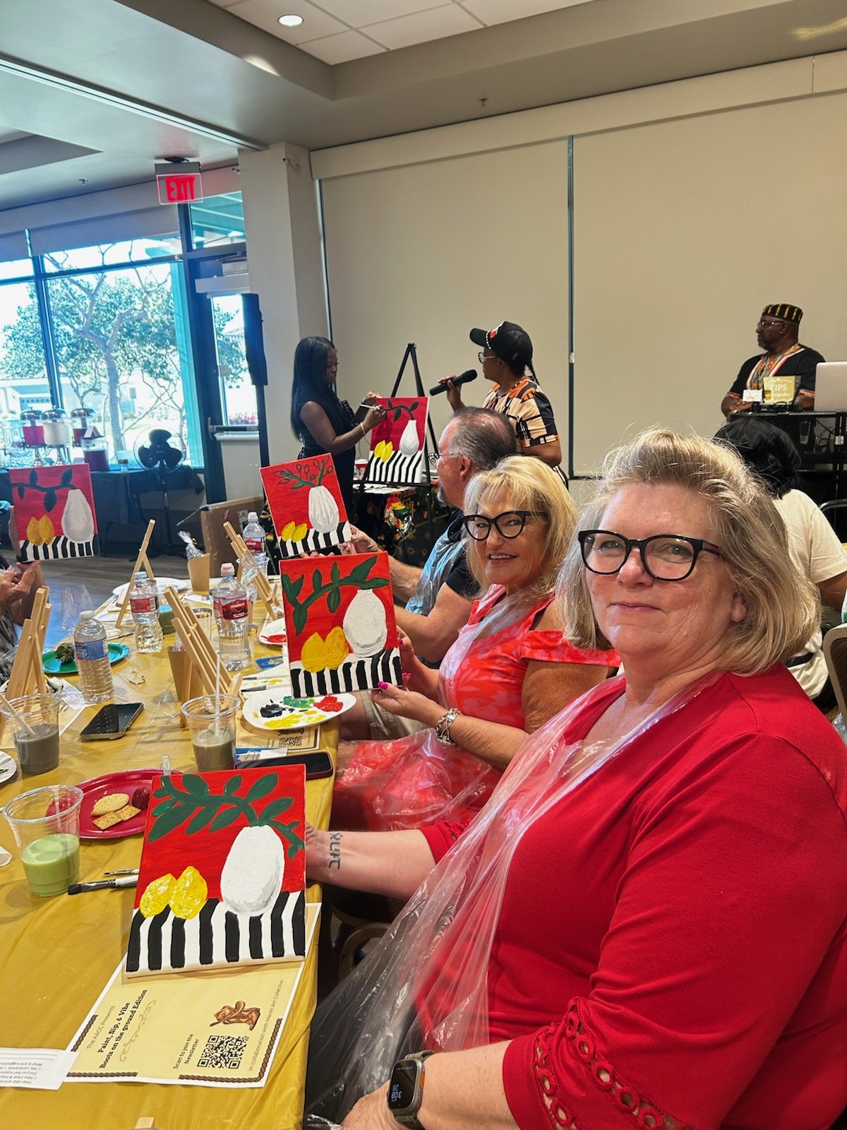Group of women participating in a painting activity at a table, with completed paintings of abstract vases and flowers in bright colors, in a lively indoor setting with large windows and a woman speaking into a microphone.