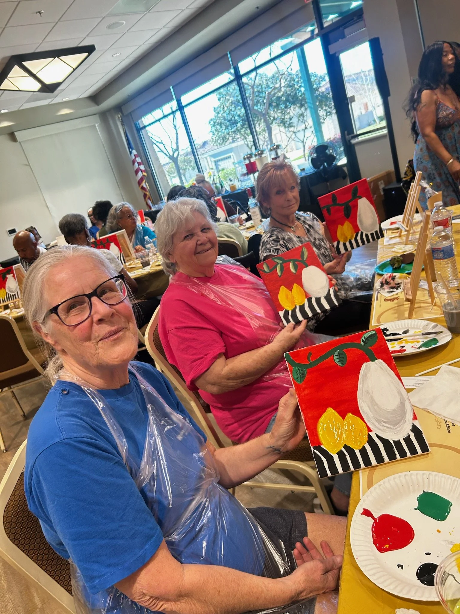 A group of older women at a painting party, holding up their finished artwork of festive holiday paintings with a red background, white ornaments, yellow accents, and black and white striped patterns, seated at tables with painting supplies.