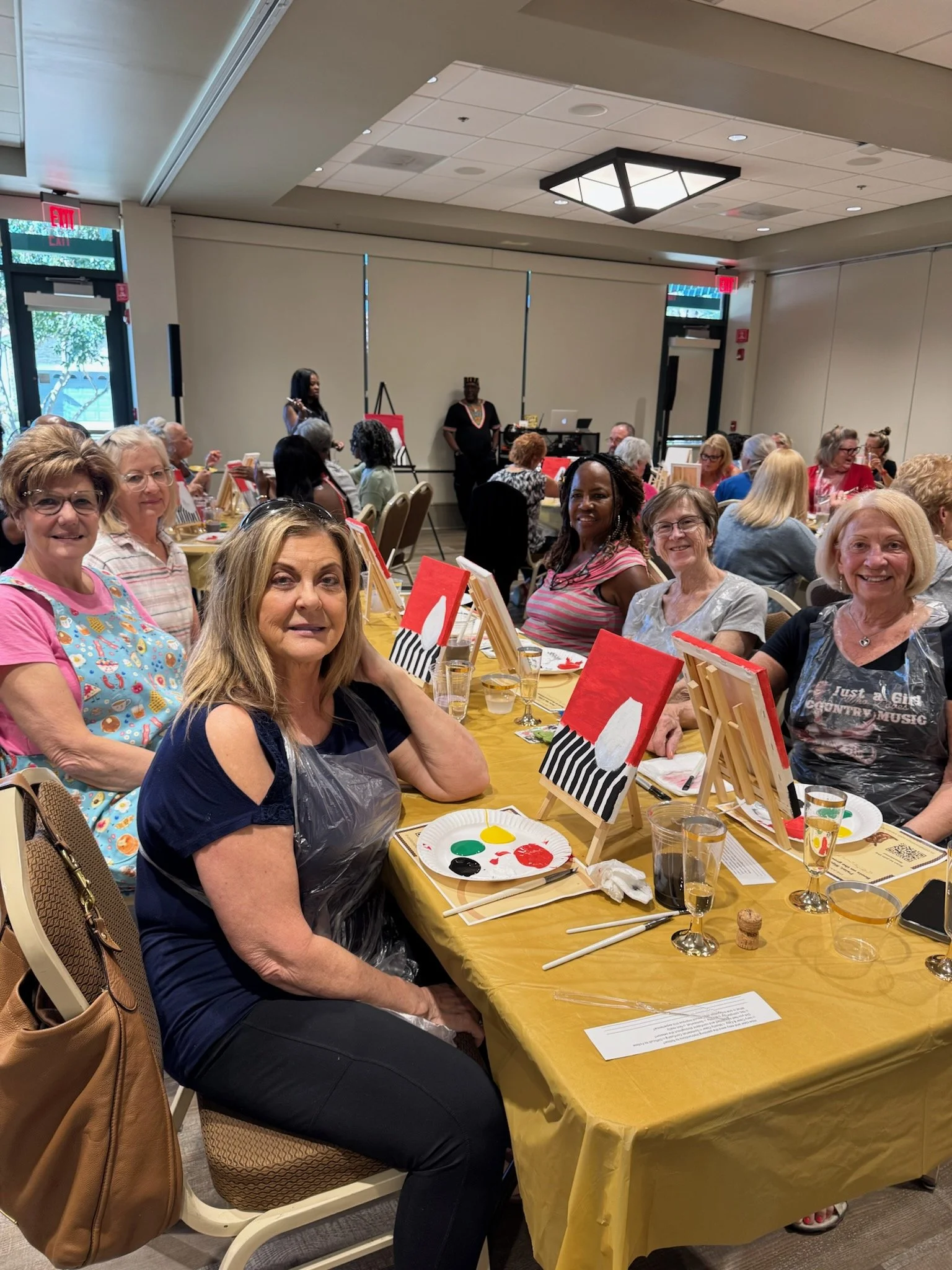 A group of women seated at a table during a paint and sip event, with canvas and paints in front of them, in a large indoor event space.