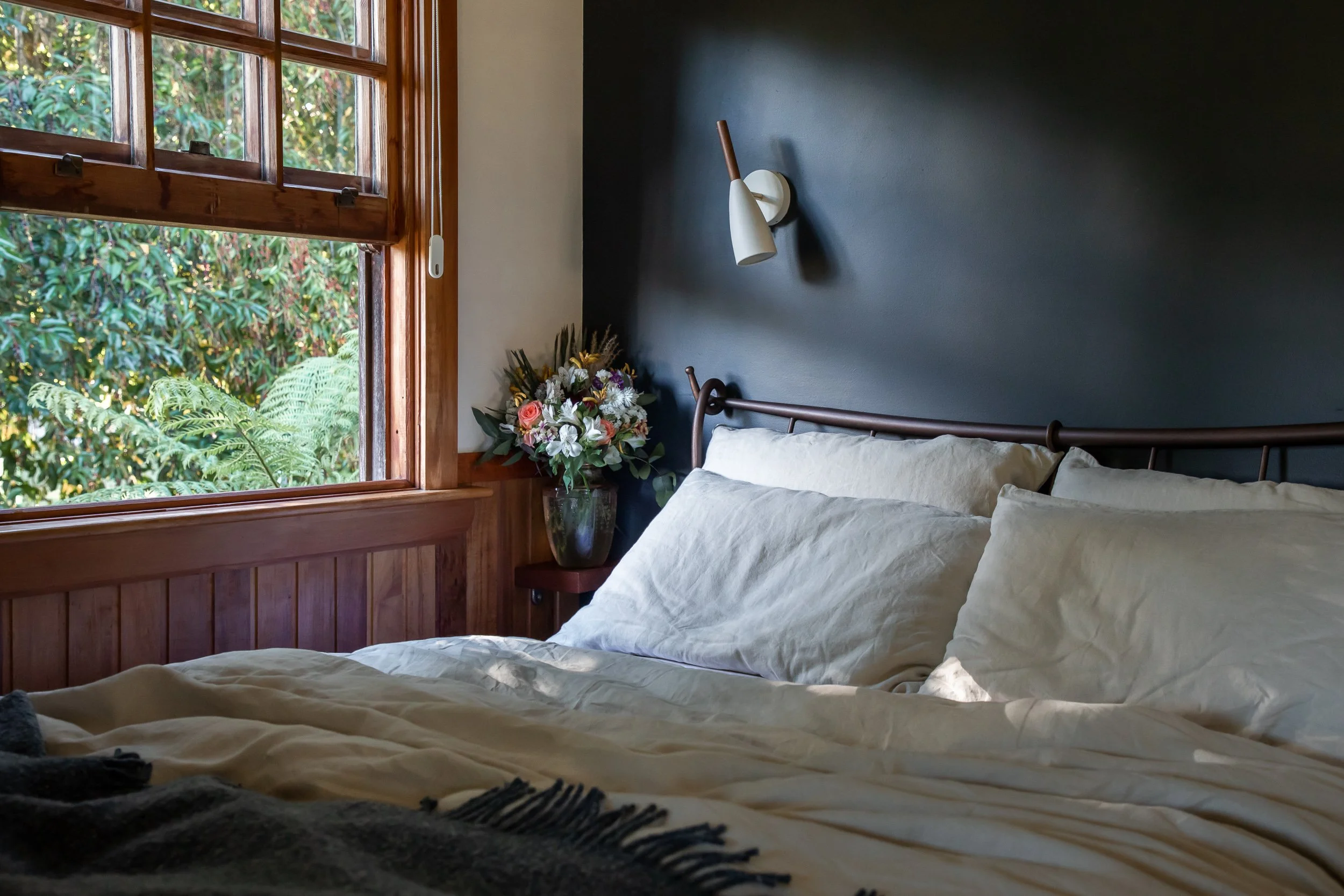 Cozy bedroom with a comfortable bed against a dark blue feature wall. The bed is covered with linen bedding and a woollen blanket. An open window shows green foliage and tree ferns outside. A vase of flowers adds colour on the bedside table.
