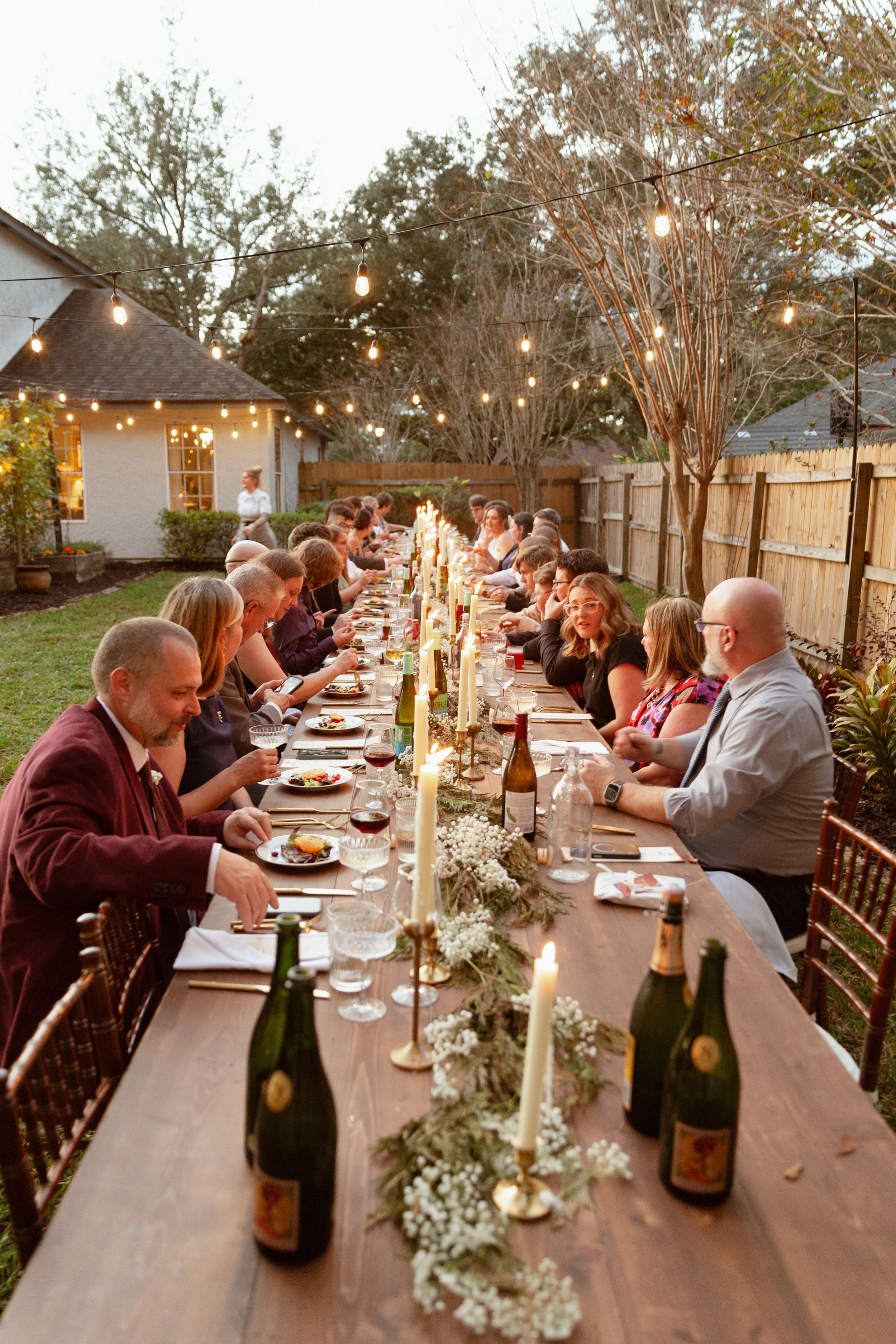 An outdoor Backyard Micro Wedding in St. Augustine, Florida. Includes a beautifully decorated Long outdoor dinner table with candles and wine bottles, guests eating and talking, string lights above, backyard with trees and house in the background.