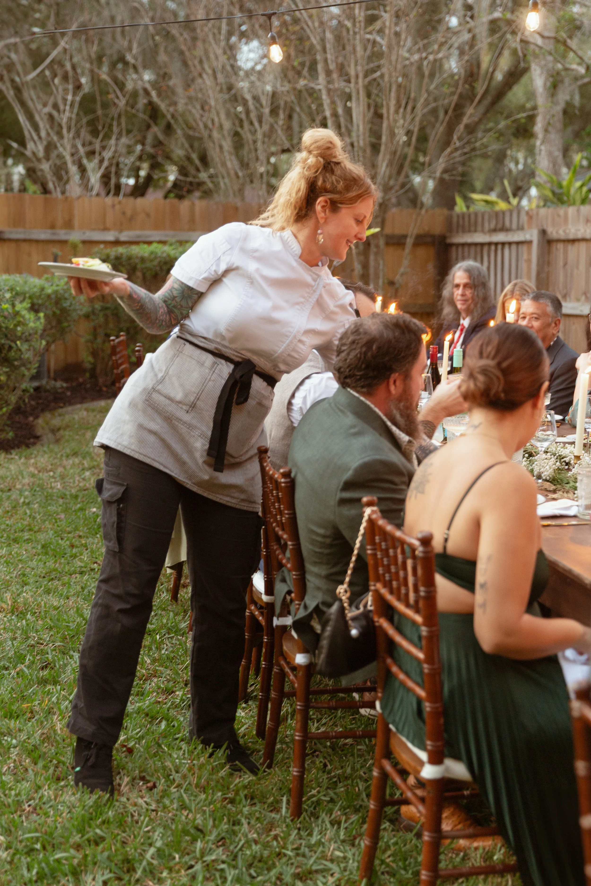 A woman with red hair styled in a bun, wearing a white shirt and an apron, is serving food to guests seated at a long outdoor table during a gathering in a backyard, with string lights hanging above and trees in the background.