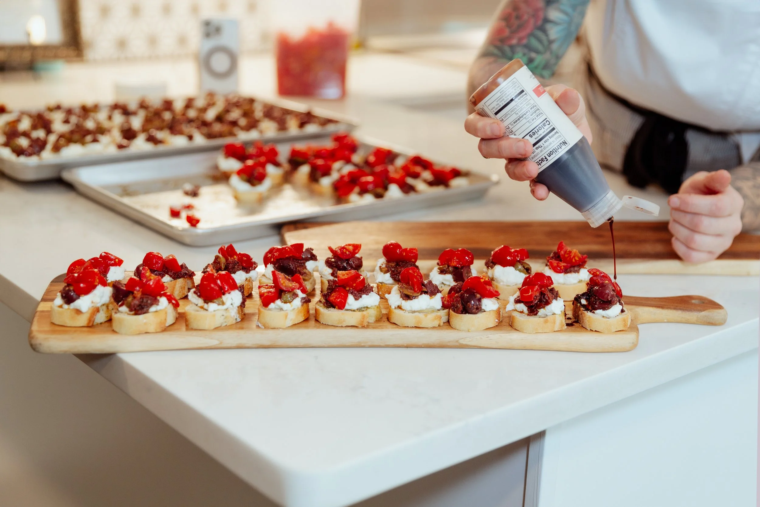 A person in a white shirt and tattooed arm is squeezing chocolate syrup onto small snacks topped with whipped cream, cherry, and fruit pieces on a wooden tray, with additional snacks on baking sheets in the background.