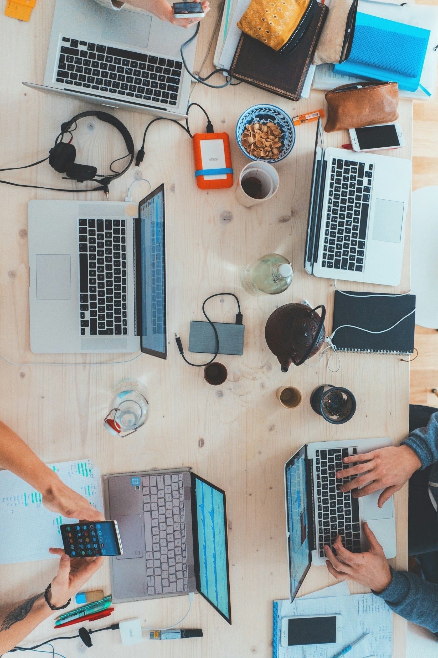 Bovenaanzicht van een houten tafel met laptops, smartphones, kabels, notitieboekjes, pennen, een kopje koffie, een waterfles, snacks, en mensen die met apparaten werken.