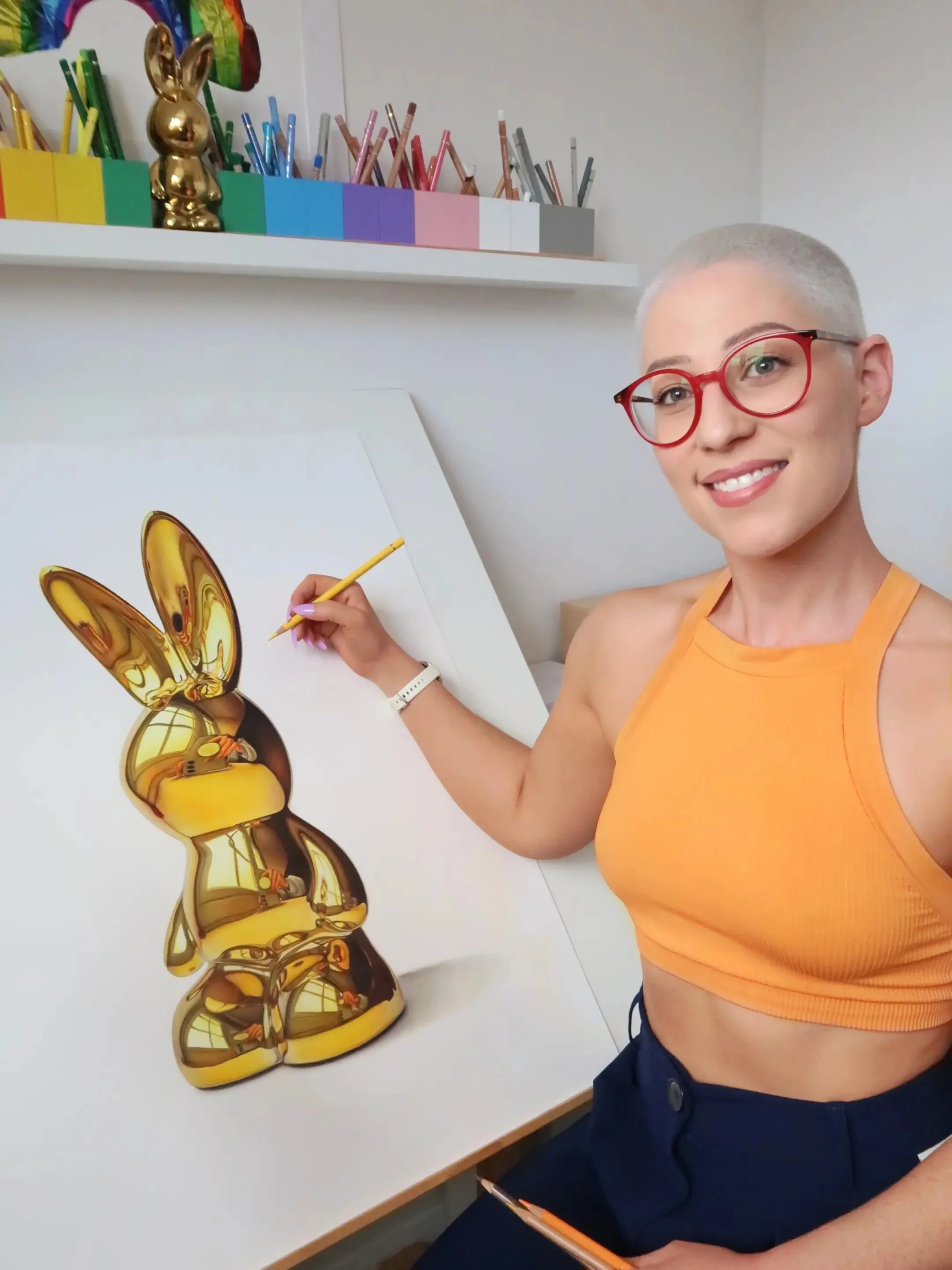 Artist Hannah Lipsey, a young women with short white hair and red glasses, holding a coloured pencil over a drawing of a gold bunny on an angled easel. Pots of coloured pencils are lined up on a shelf above her.