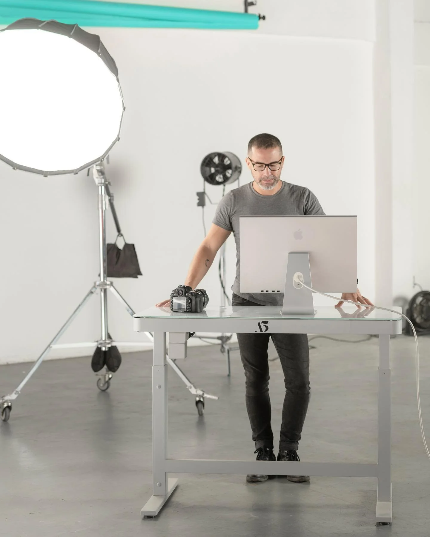 Hombre con gafas en un estudio fotográfico, trabajando frente a una computadora en una mesa de color blanco, con equipo de iluminación, un reflector y una cámara en la mesa.