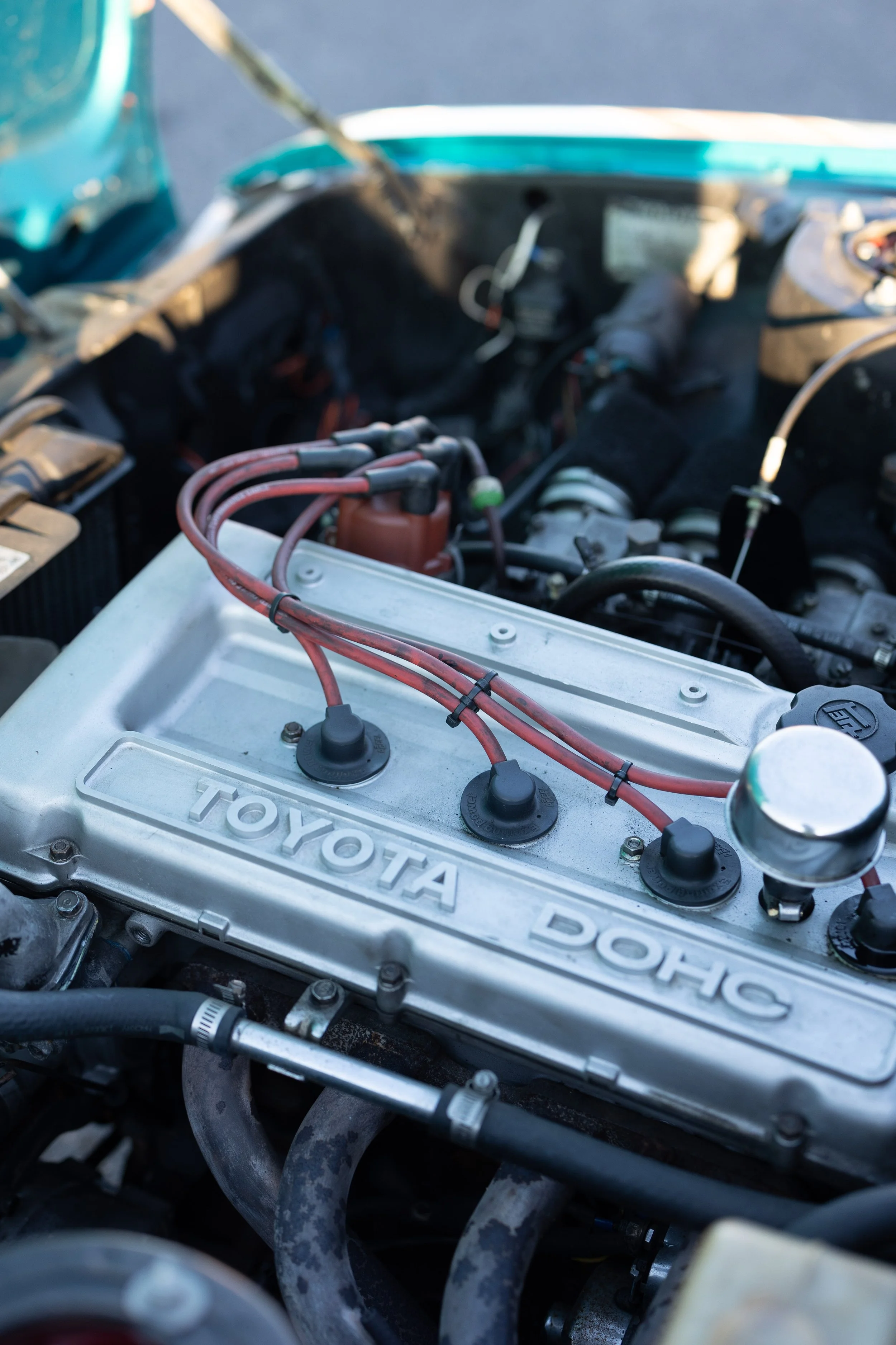 Toyota 2T-G DOHC twin-cam engine in a Celica 1600GT TA22 with red ignition leads and an alloy cam cover. Classic Japanese engine bay photography.