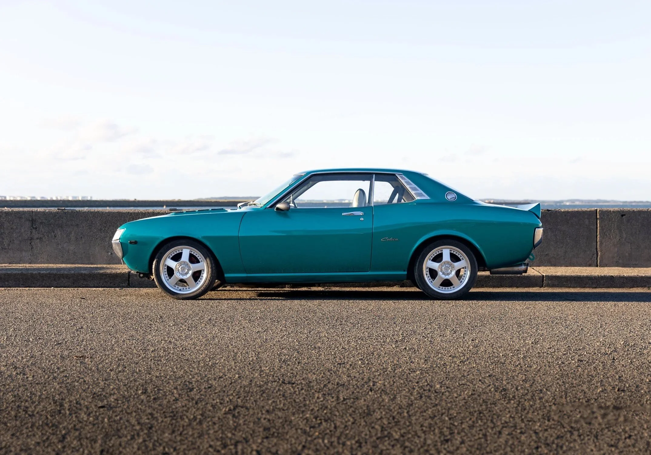 Toyota Celica 1600GT TA22 full side profile in teal metallic, lowered on polished deep-dish alloys by the water classic JDM coupe car photography.