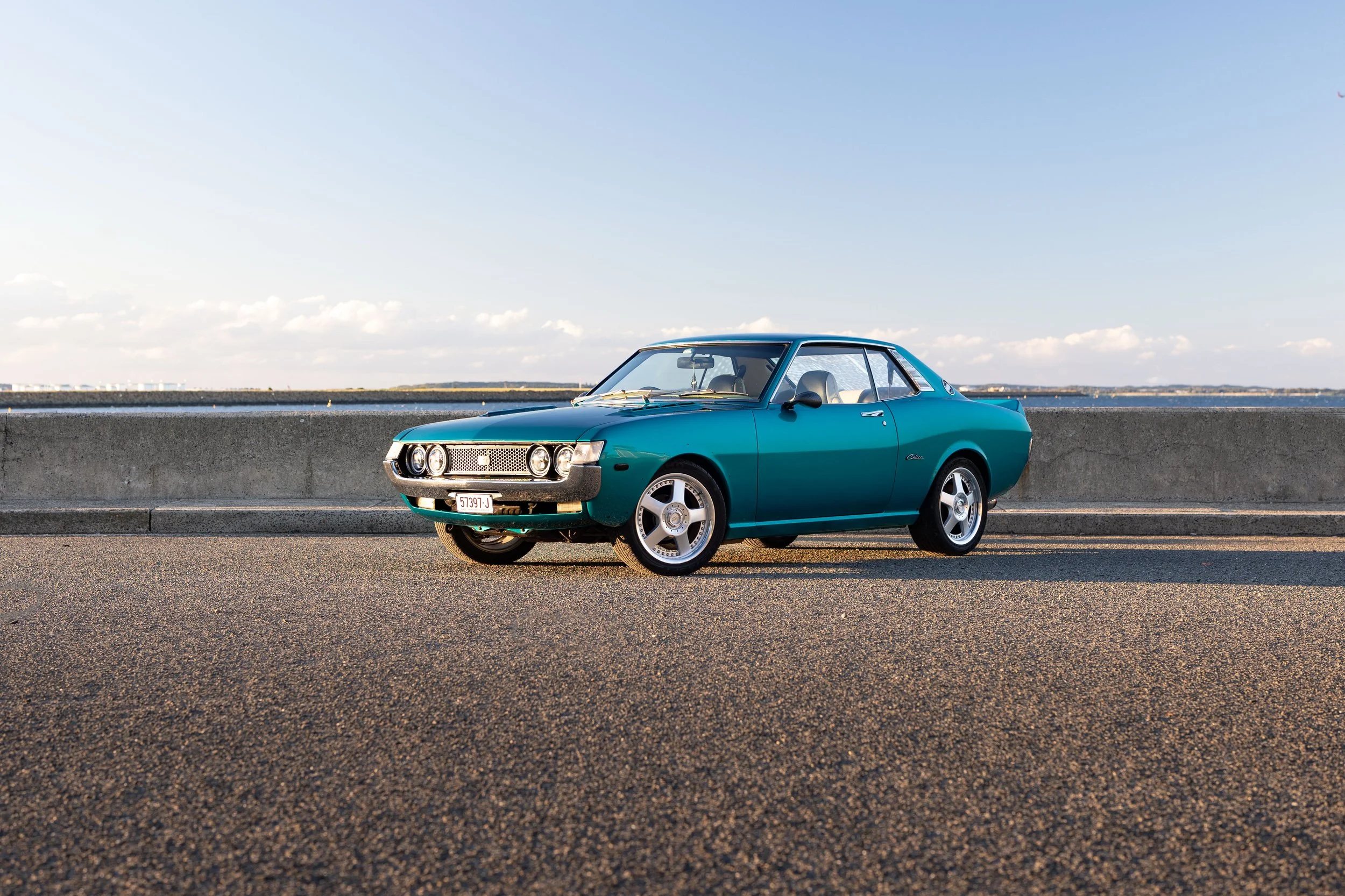 Toyota Celica 1600GT TA22 front three-quarter low-angle by the water, teal coupe on staggered deep-dish wheels classic Japanese car photography.