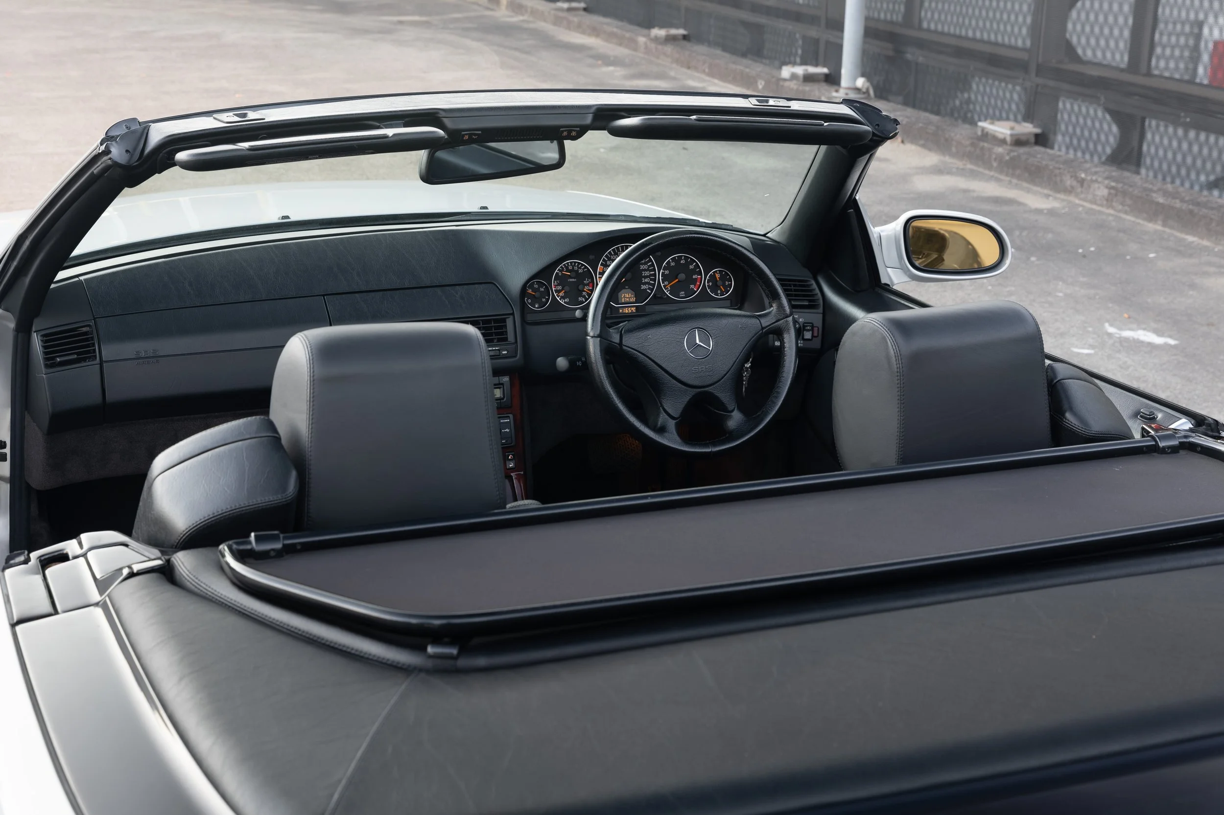Interior of a silver R129 Mercedes-Benz convertible car showing the dashboard, steering wheel, and front seats, with the roof down.