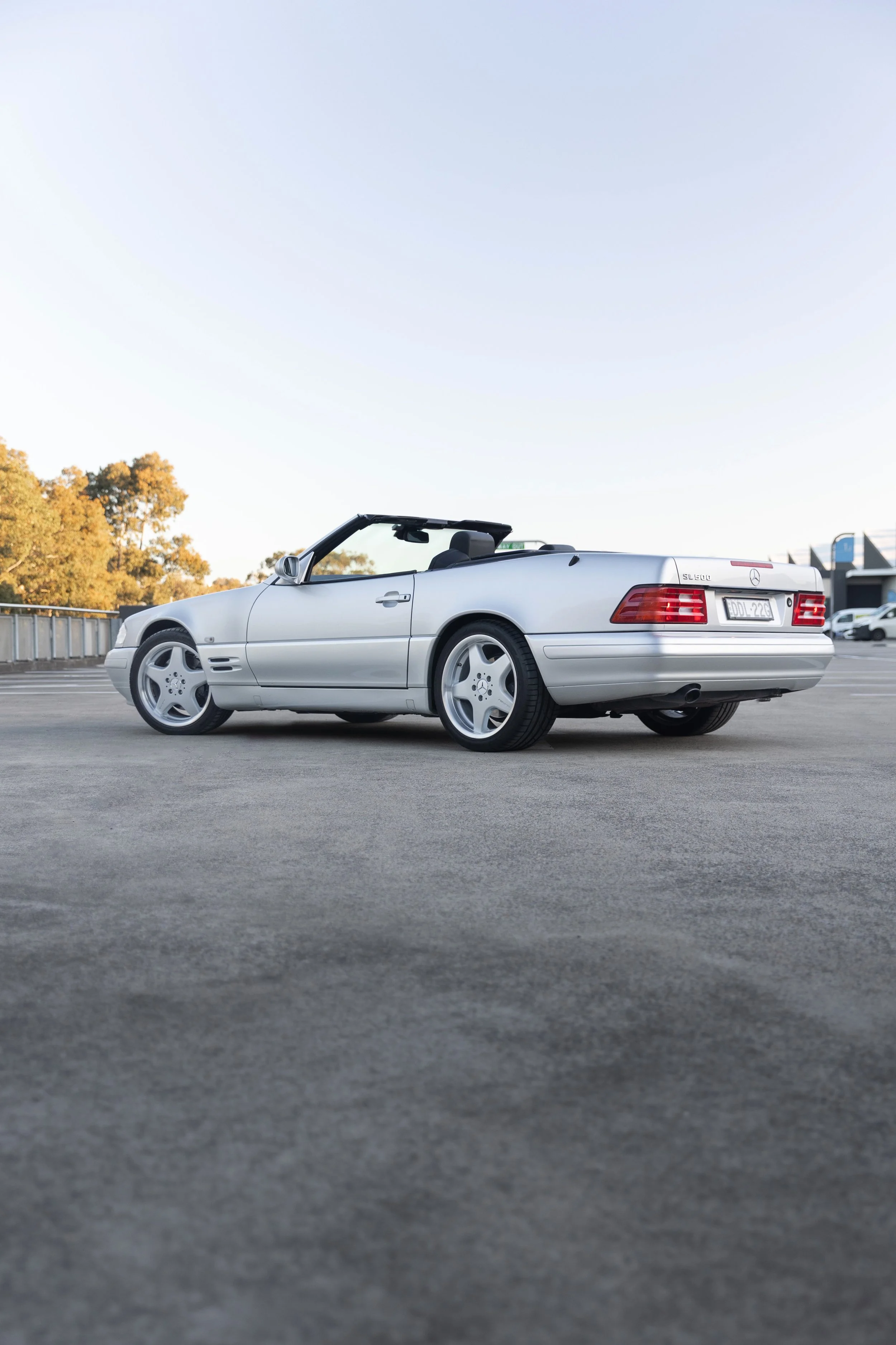 1999 Mercedes-Benz SL 500 R129 rear three-quarter on a Sydney rooftop, leafy backdrop, roof down.