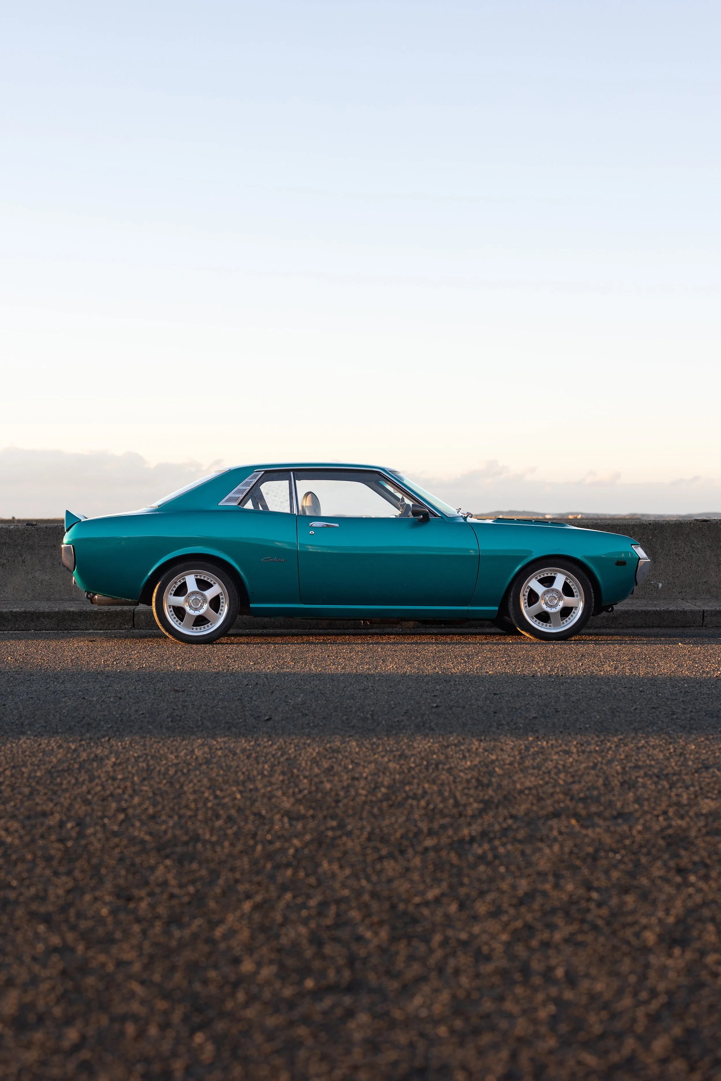 Toyota Celica 1600GT TA22 driver-side profile at golden hour on a harbour seawall, long-bonnet classic coupe silhouette automotive photography.