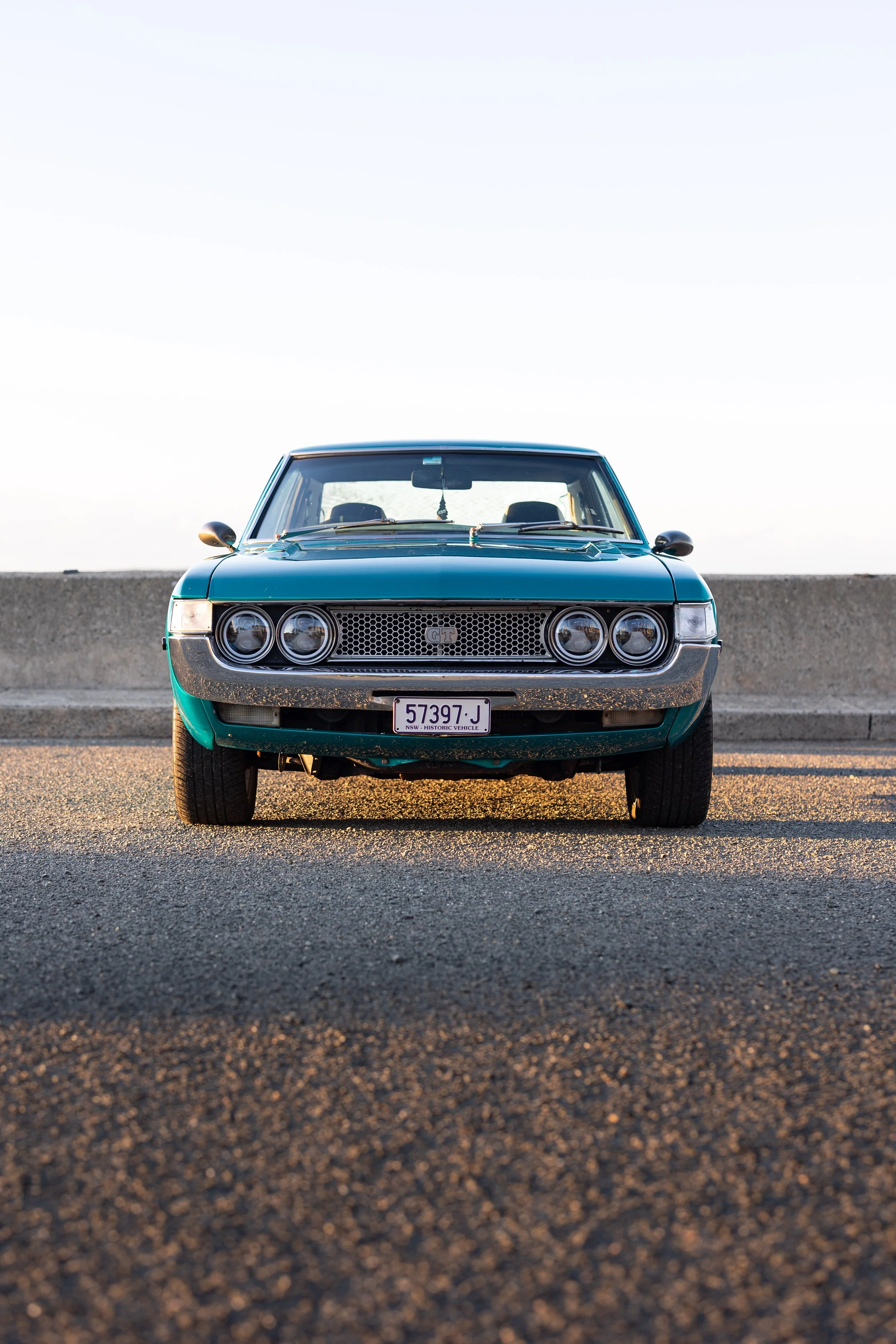 Toyota Celica 1600GT TA22 head-on at a coastal seawall, twin round headlights, mesh GT grille and NSW historic plate on teal paint classic Japanese coupe automotive photography.